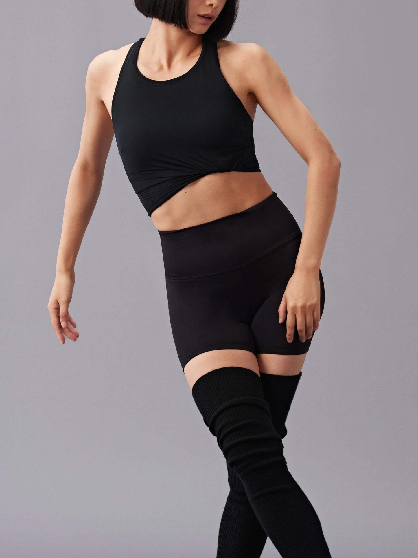 A woman wearing a black sports bra top, high-waisted black shorts, and black leg warmers poses against a plain gray background.