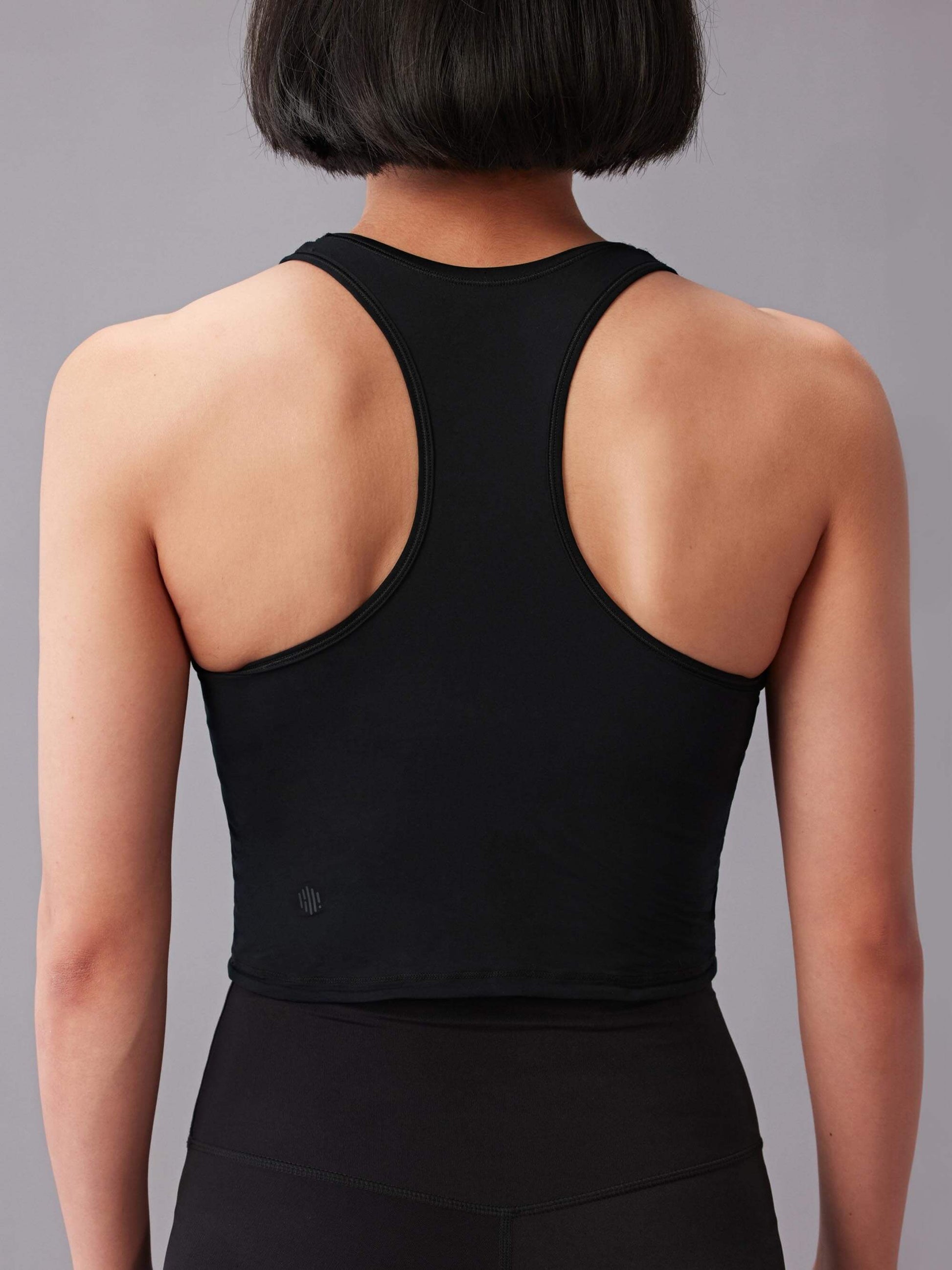 The image depicts the back view of a person wearing a black racerback sports bra top. The top has a close fit and the fabric appears smooth and lightweight. 