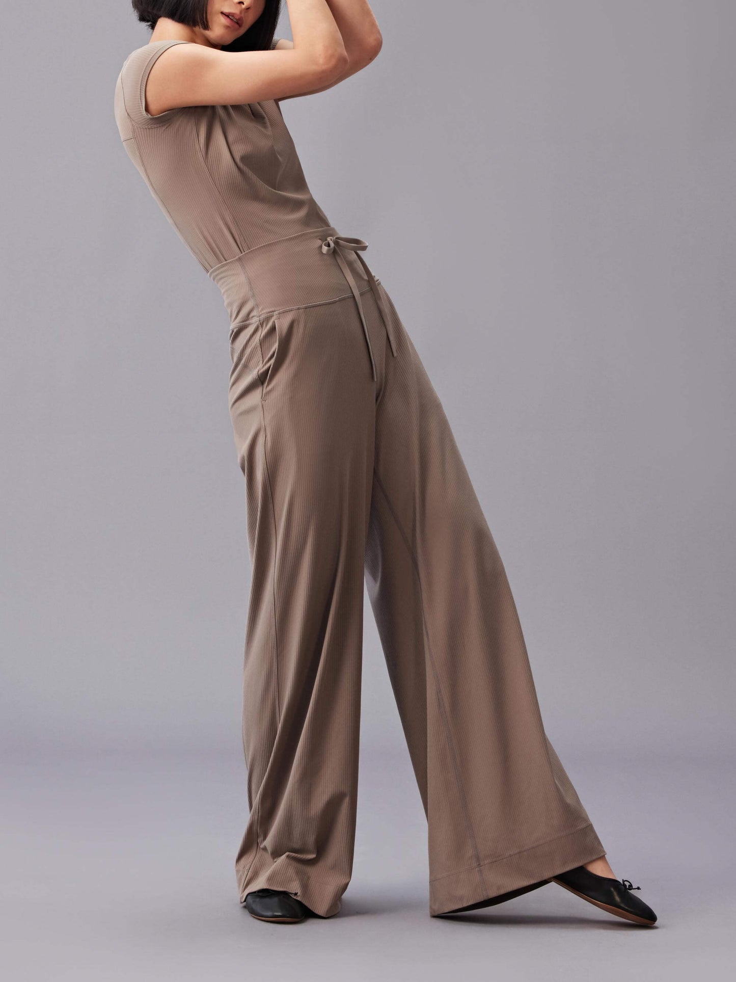 A woman wearing a beige, short-sleeve top and matching high-waisted, wide-leg pants, posing with arms raised and bent, against a gray background.