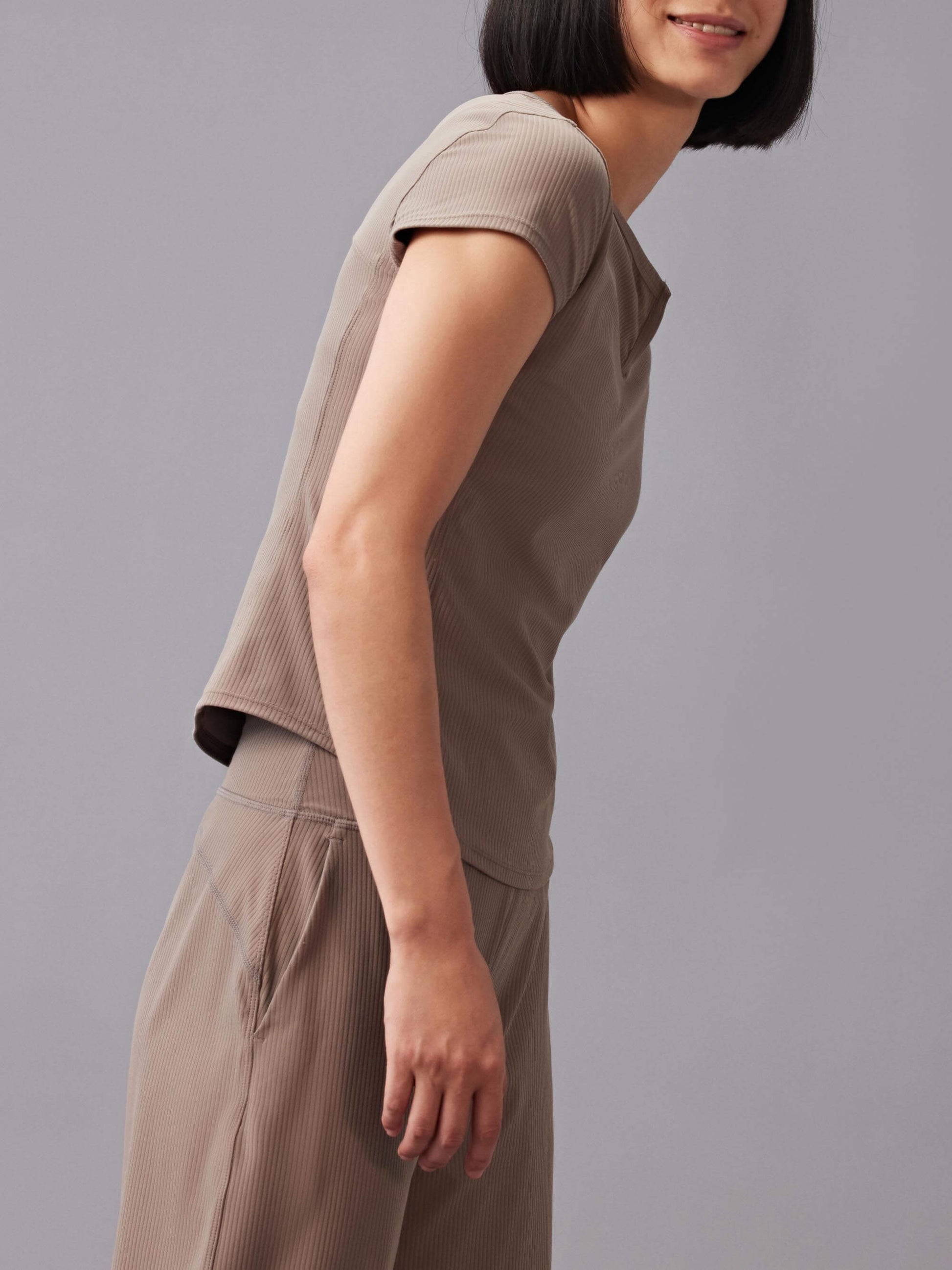 Woman wearing a taupe-colored short-sleeve top and matching pants, leaning slightly forward.