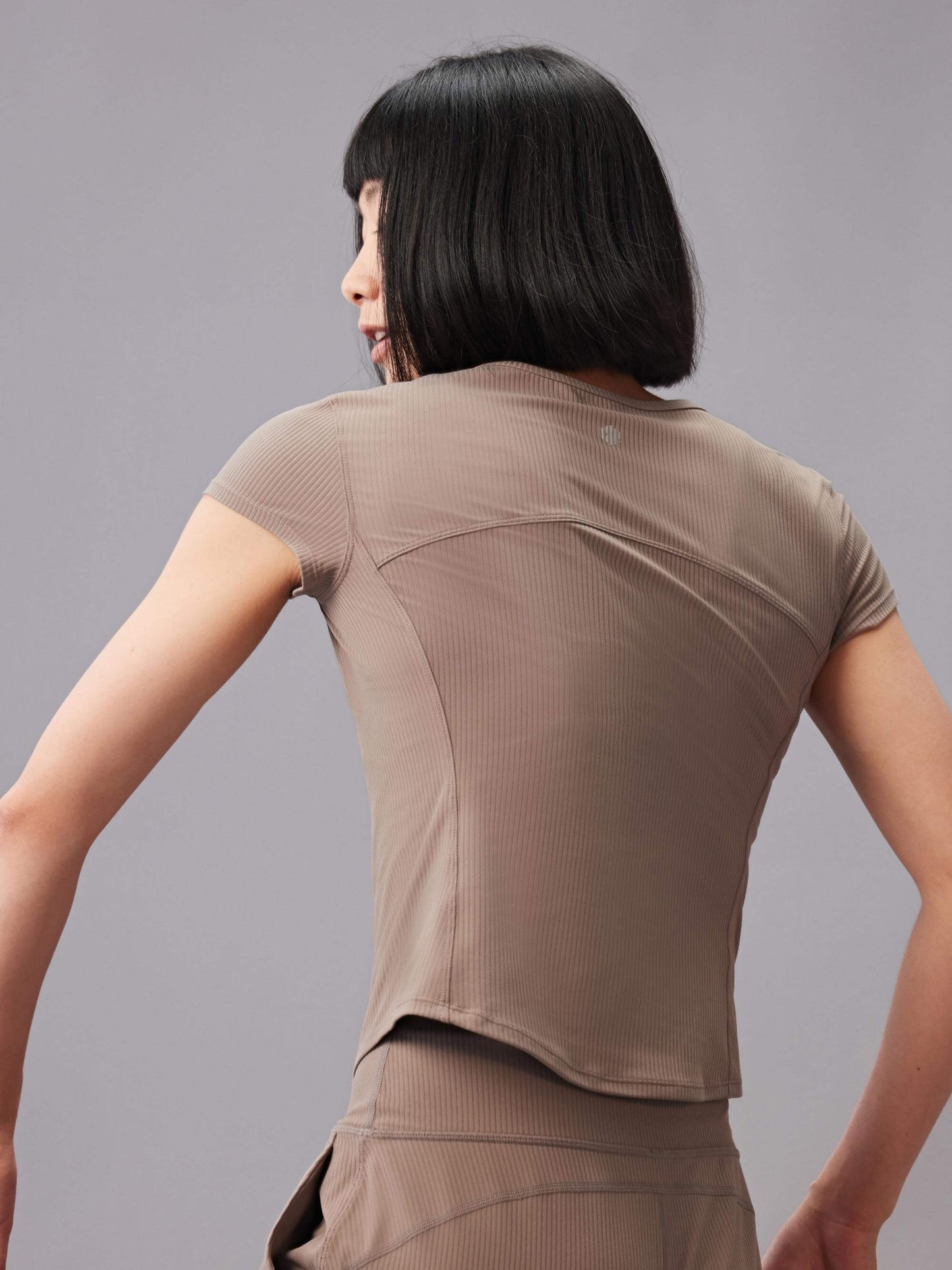 Woman in a taupe short-sleeve top and matching pants, seen from the back, turning her head to the side.