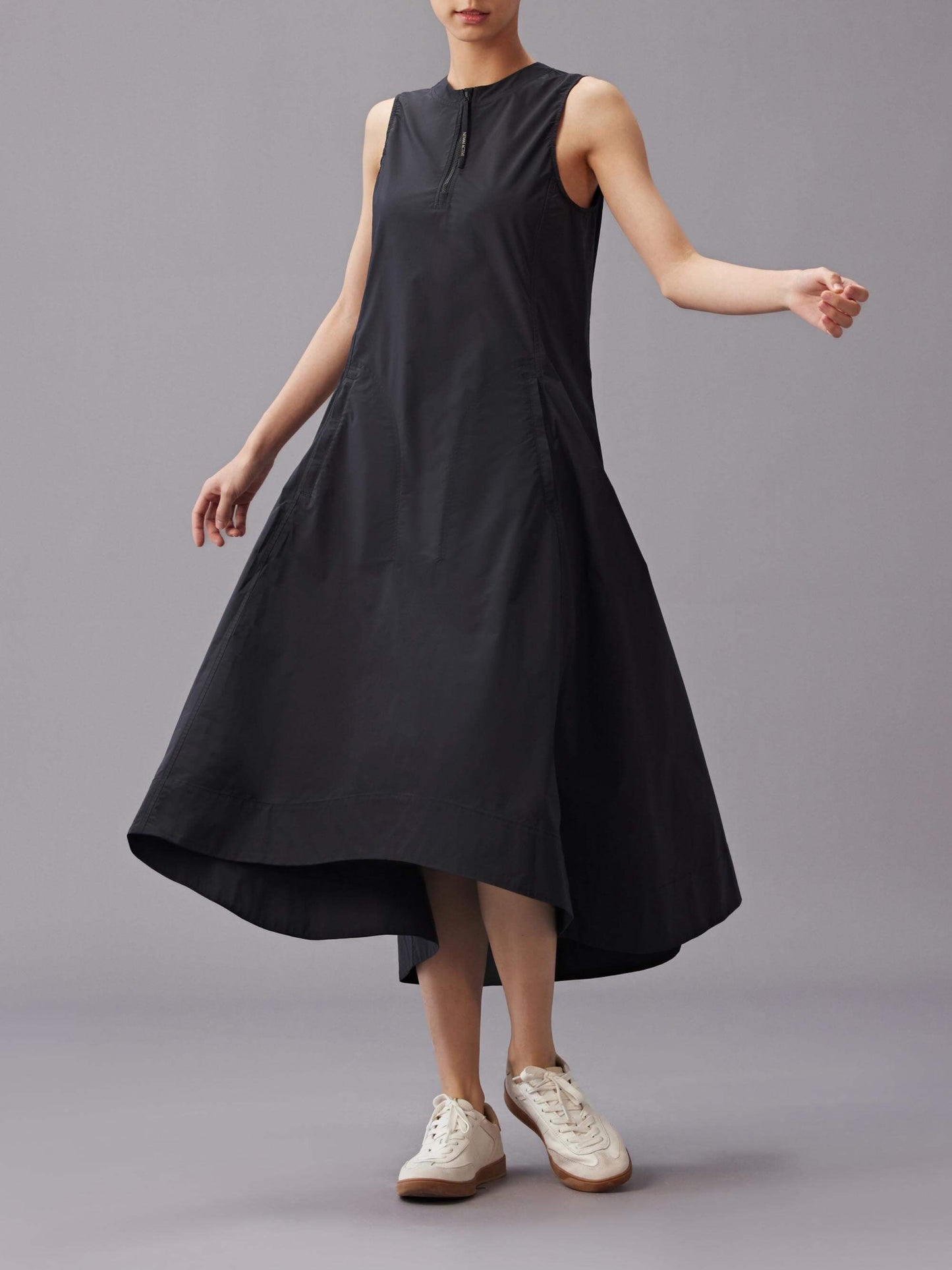 Model wearing a sleeveless black dress with an asymmetrical hemline, paired with white sneakers. The dress features a front zipper detail and a flowy, A-line silhouette.