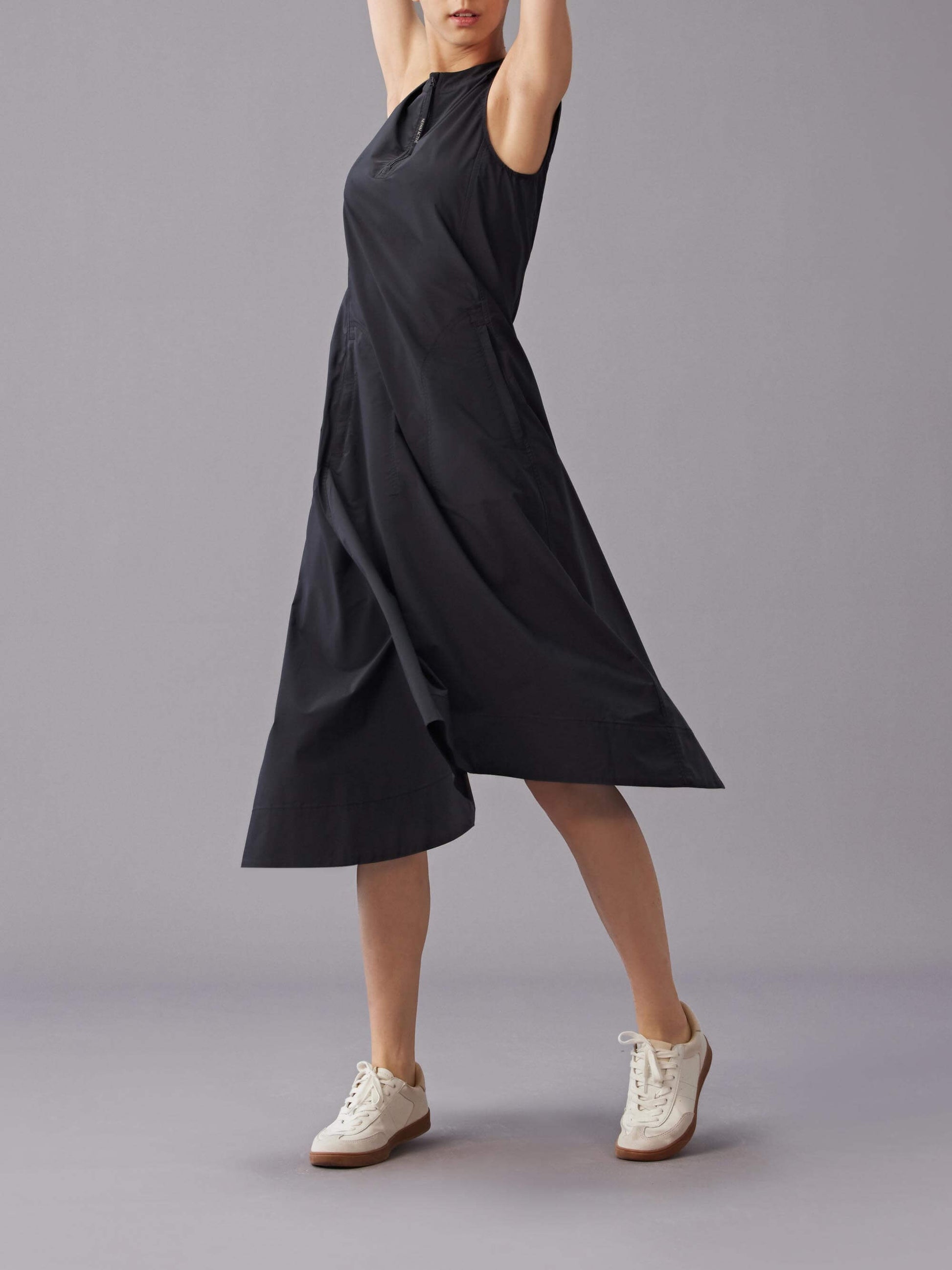 Model wearing a sleeveless black dress with an asymmetrical hemline, striking a pose with arms raised, paired with white sneakers.