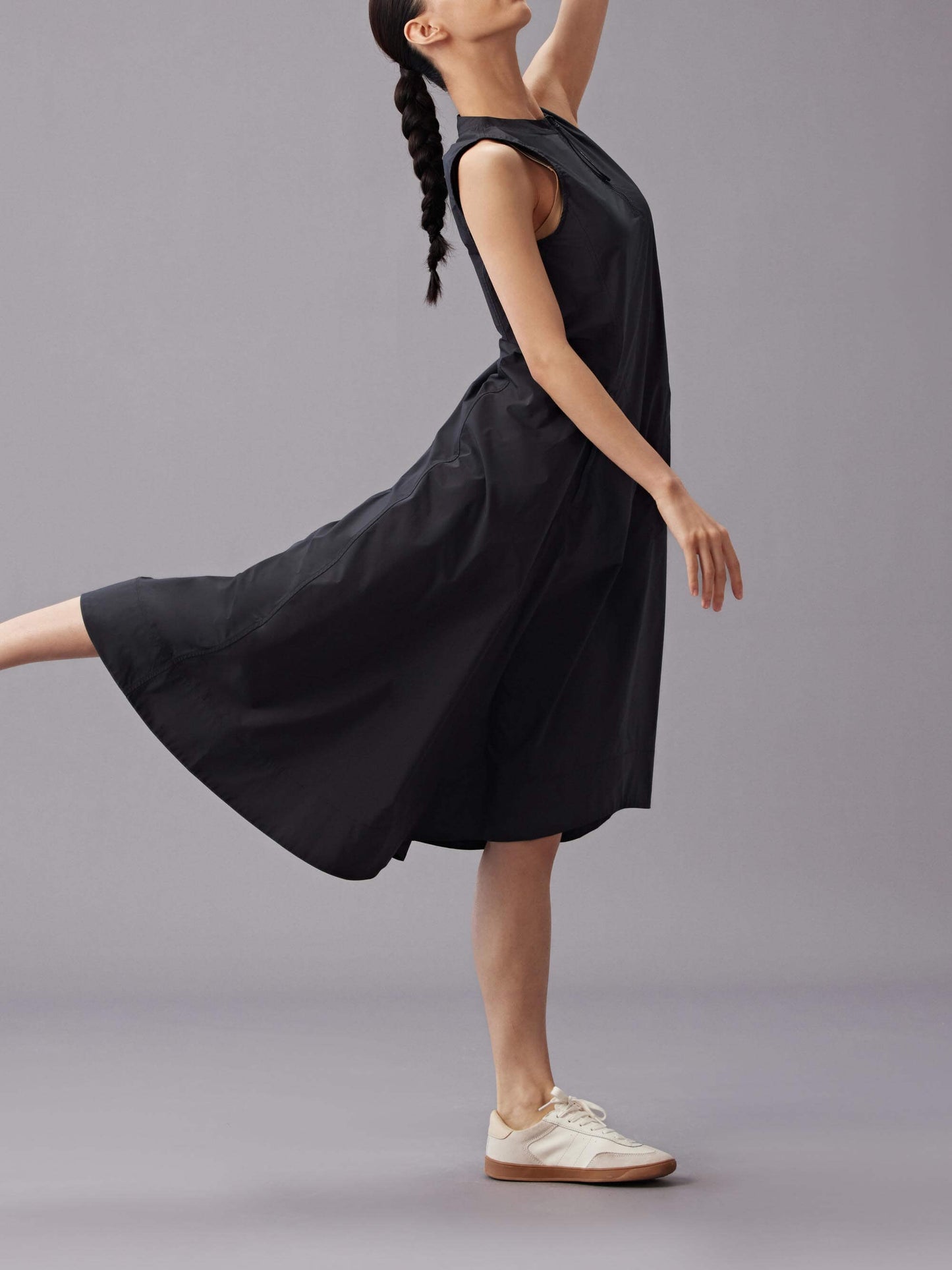 Model wearing a sleeveless black dress with an asymmetrical hemline, gracefully posing on one leg, paired with white sneakers.