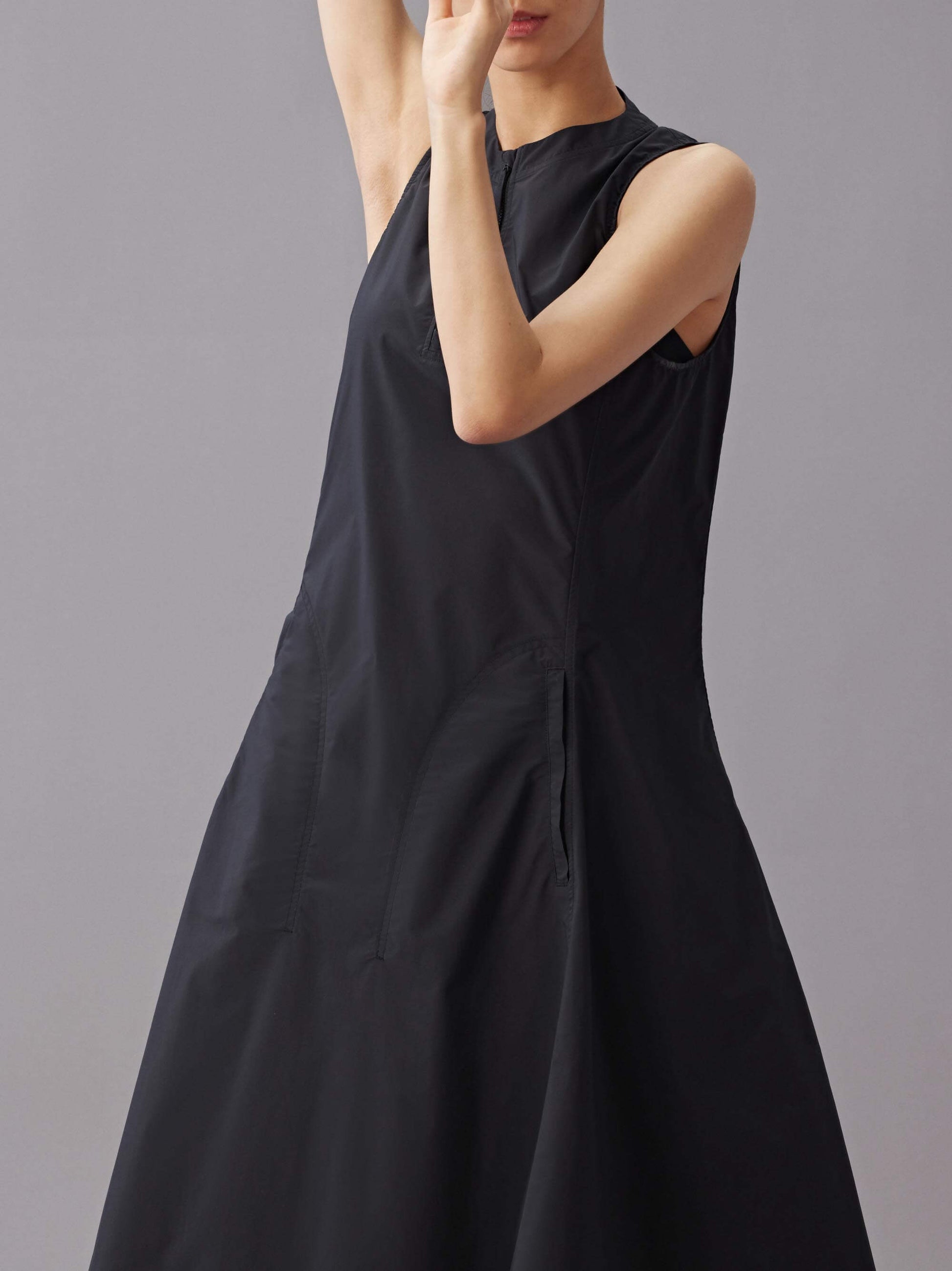 Model in a close-up shot wearing a sleeveless black dress with an A-line silhouette and front zipper detail, posing with arms raised against a neutral background.