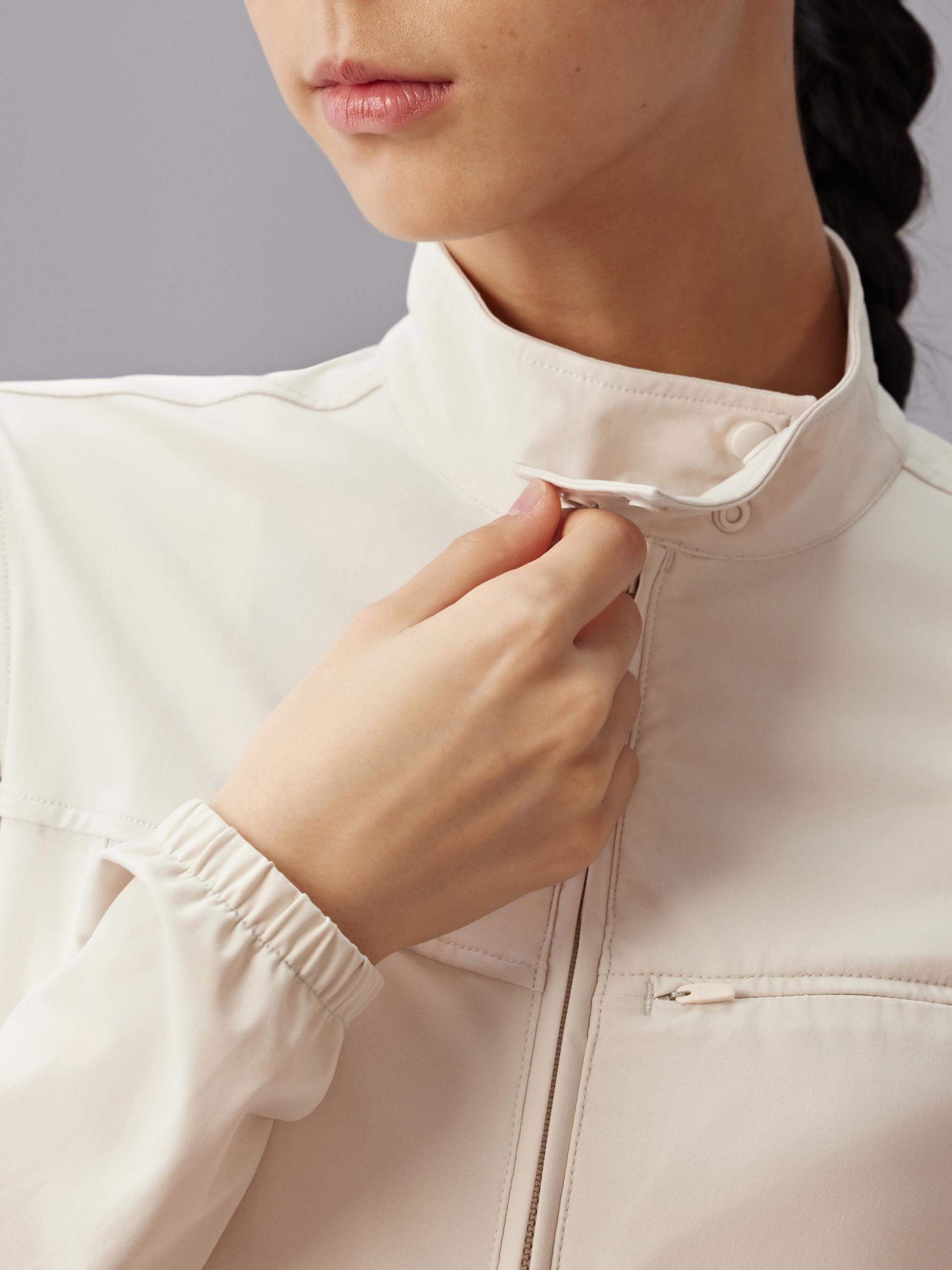 Close-up of a woman adjusting the collar of a beige fitted zip-up jacket, with a braid visible on the side, against a gray background.