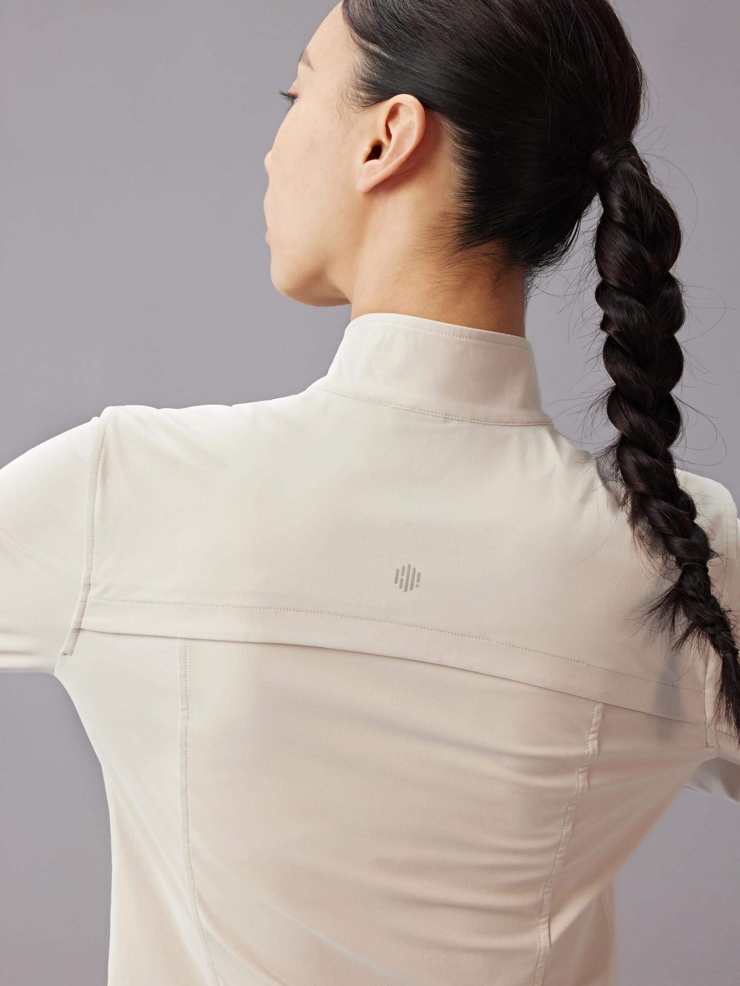 Back view of a woman with a braid, wearing a beige fitted zip-up jacket, against a gray background.