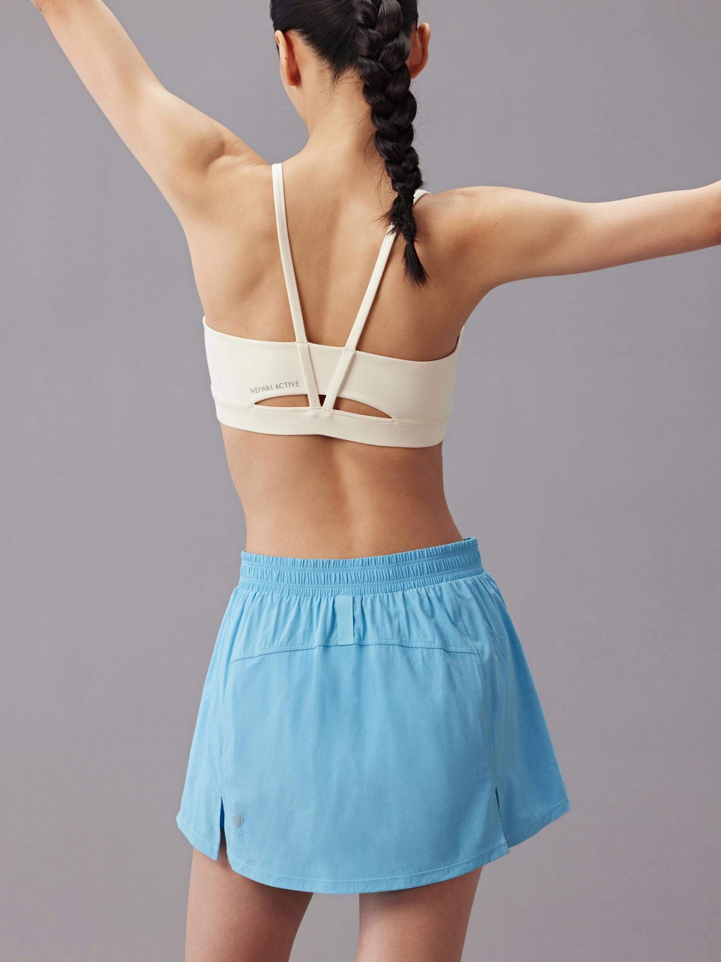 The back view of a woman wearing a white sports bra and a light blue training skort with an elastic waistband, performing a stretching pose.