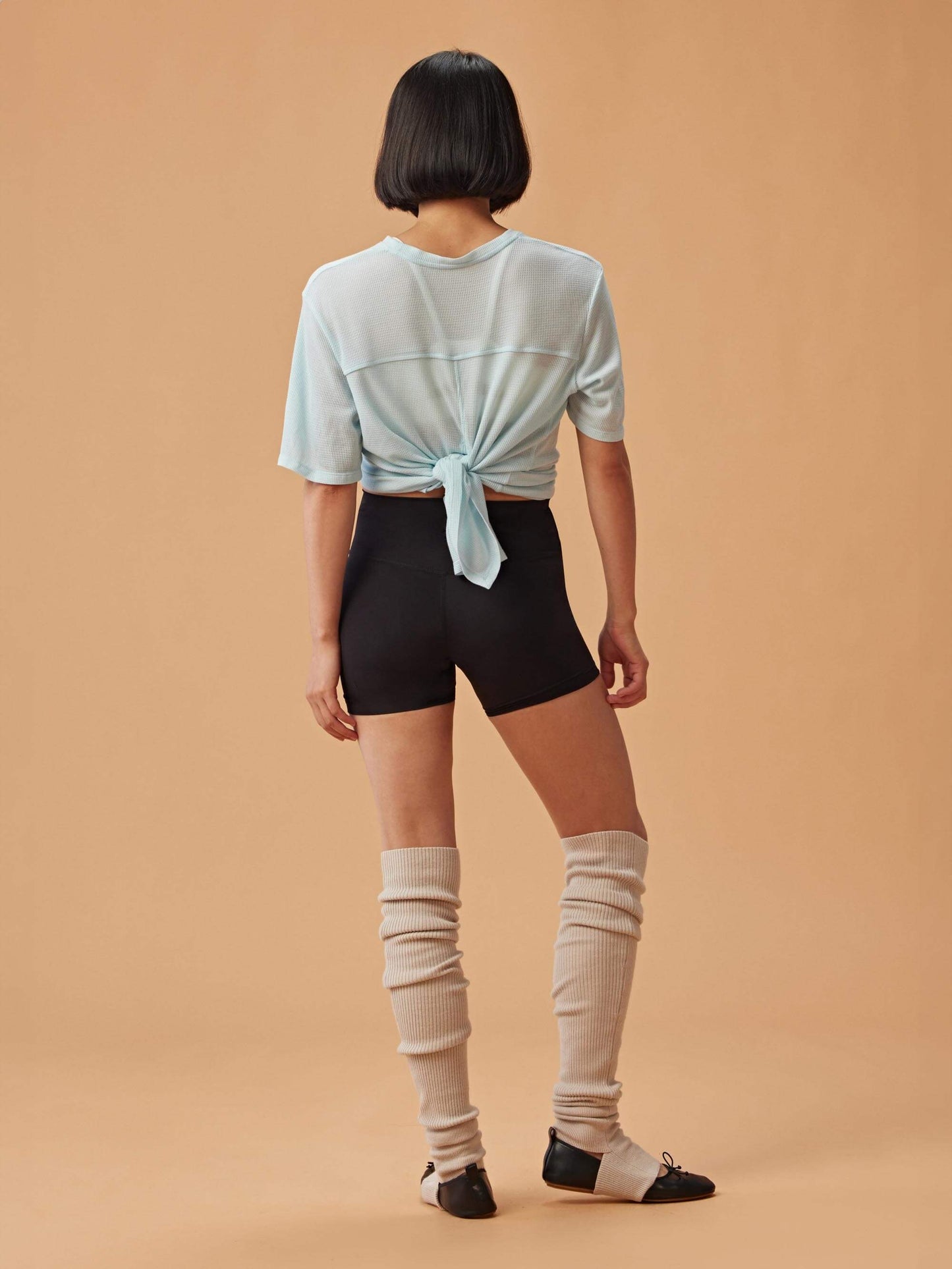 A woman wearing a light blue waffle short sleeve top with an open-back design tied in a knot, paired with black shorts and light beige leg warmers, posed with her back to the camera.