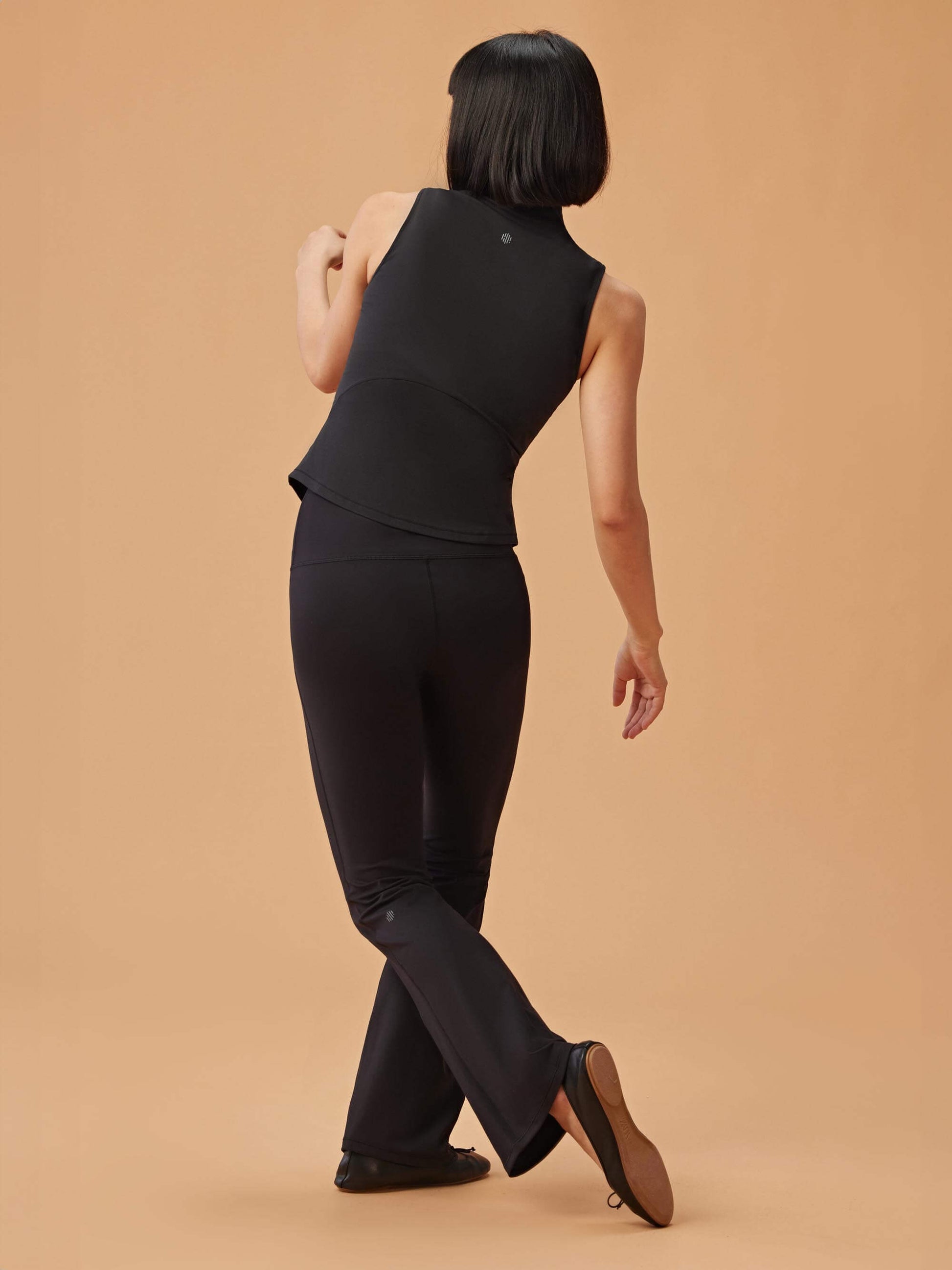 Woman wearing black high-waisted flare leggings and a black sleeveless top, standing with her back to the camera against a beige background.