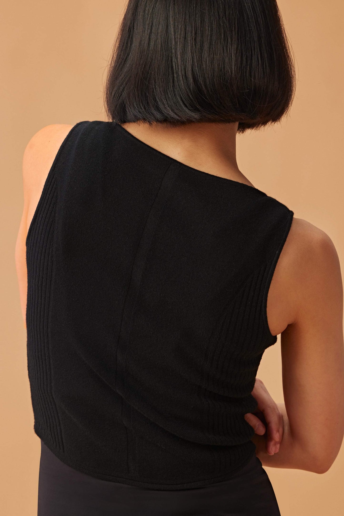 Back view of woman wearing a black sleeveless top with a ribbed lower section, viewed from the back, against a warm beige background.