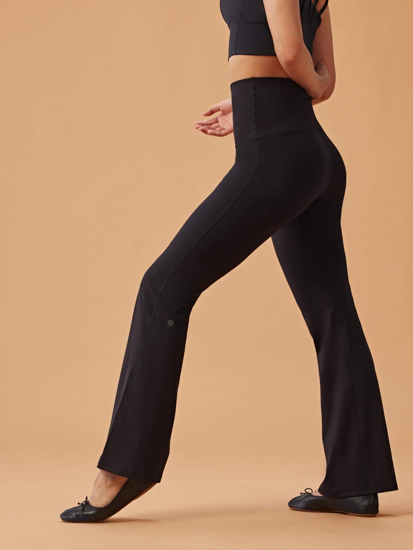 Woman wearing black high-waisted flare leggings and a black crop top, standing in profile against a beige background.