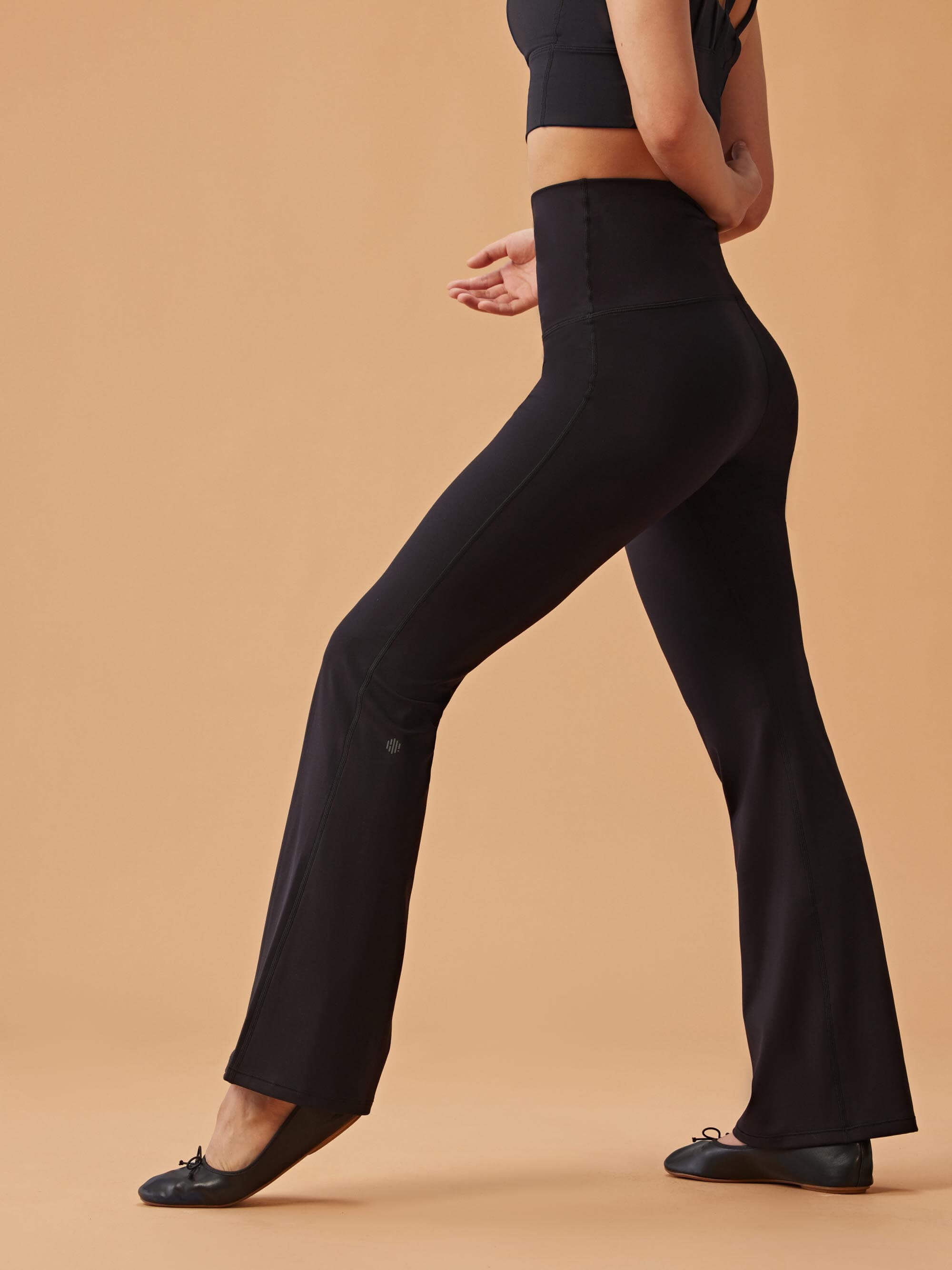 Woman wearing black high-waisted flare leggings and a black crop top, standing in profile against a beige background.