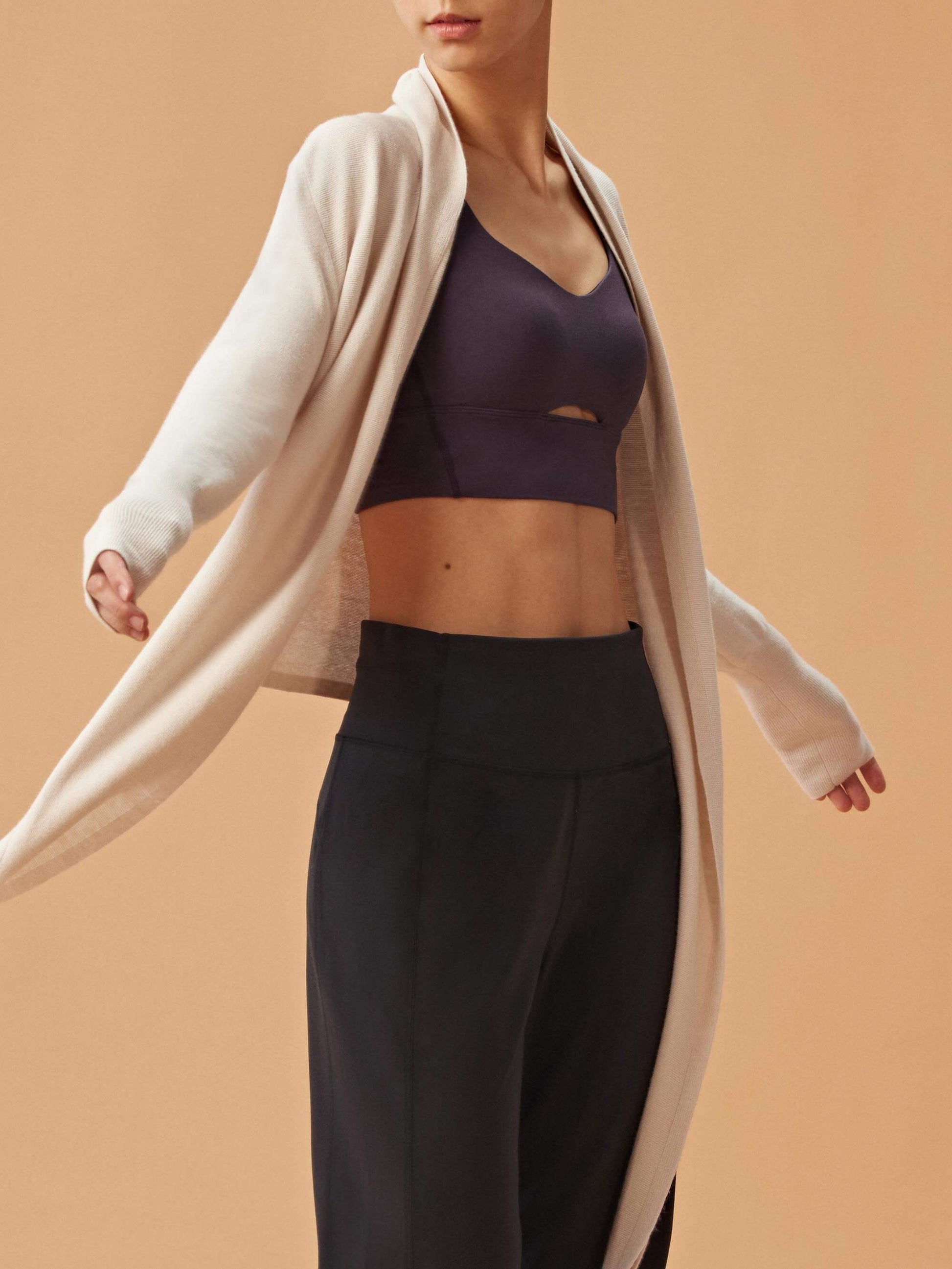 A woman wearing a light beige long cardigan over a dark sports bra and high-waisted black pants. The cardigan is open, showcasing the stylish and comfortable outfit underneath.