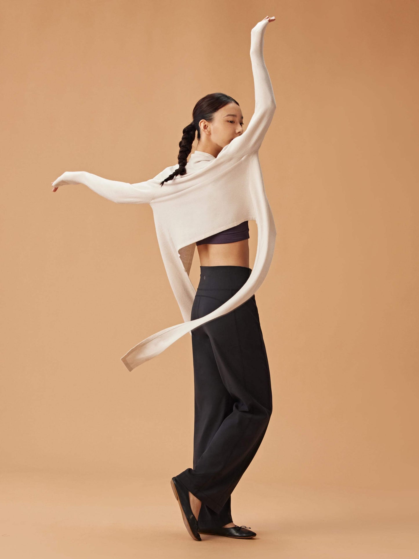 A woman dancing while wearing a beige long cardigan over a dark sports bra and high-waisted black pants. The cardigan flows behind her, creating a dynamic and elegant look.