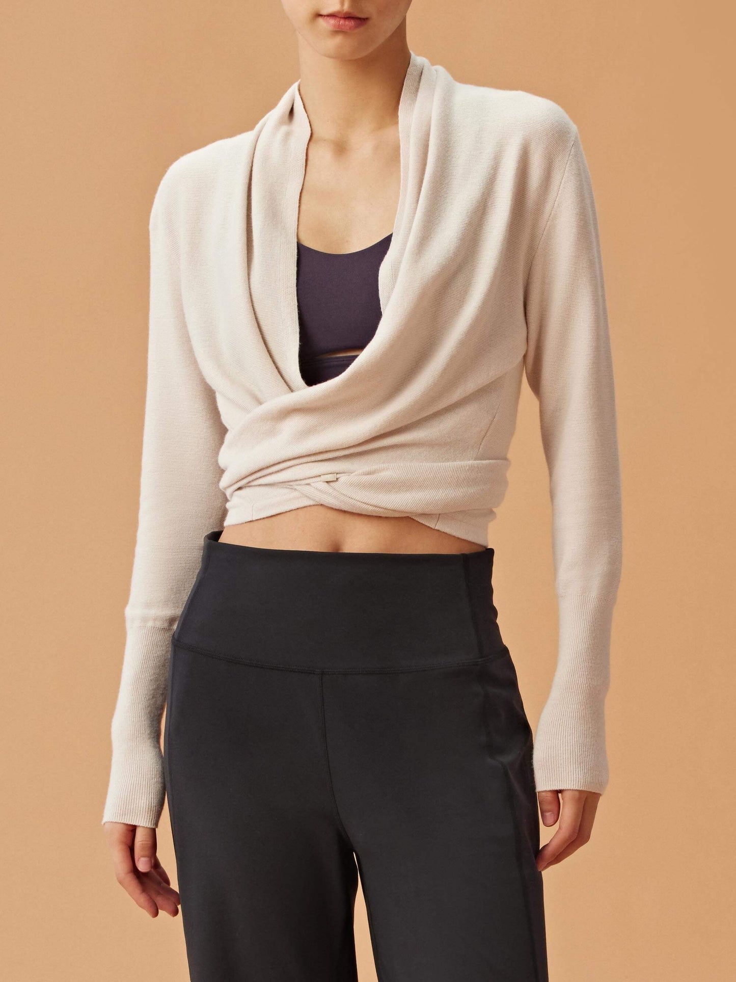 A woman wearing a cream-colored wrap-style cardigan over a dark sports bra, paired with high-waisted black pants. The cardigan has a draped front and long sleeves.