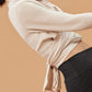 Close-up of a woman wearing a cream-colored wrap-style cardigan tied at the back, over a dark sports bra and high-waisted black pants. The cardigan has a soft, cozy texture, and the tied back detail adds a stylish touch.