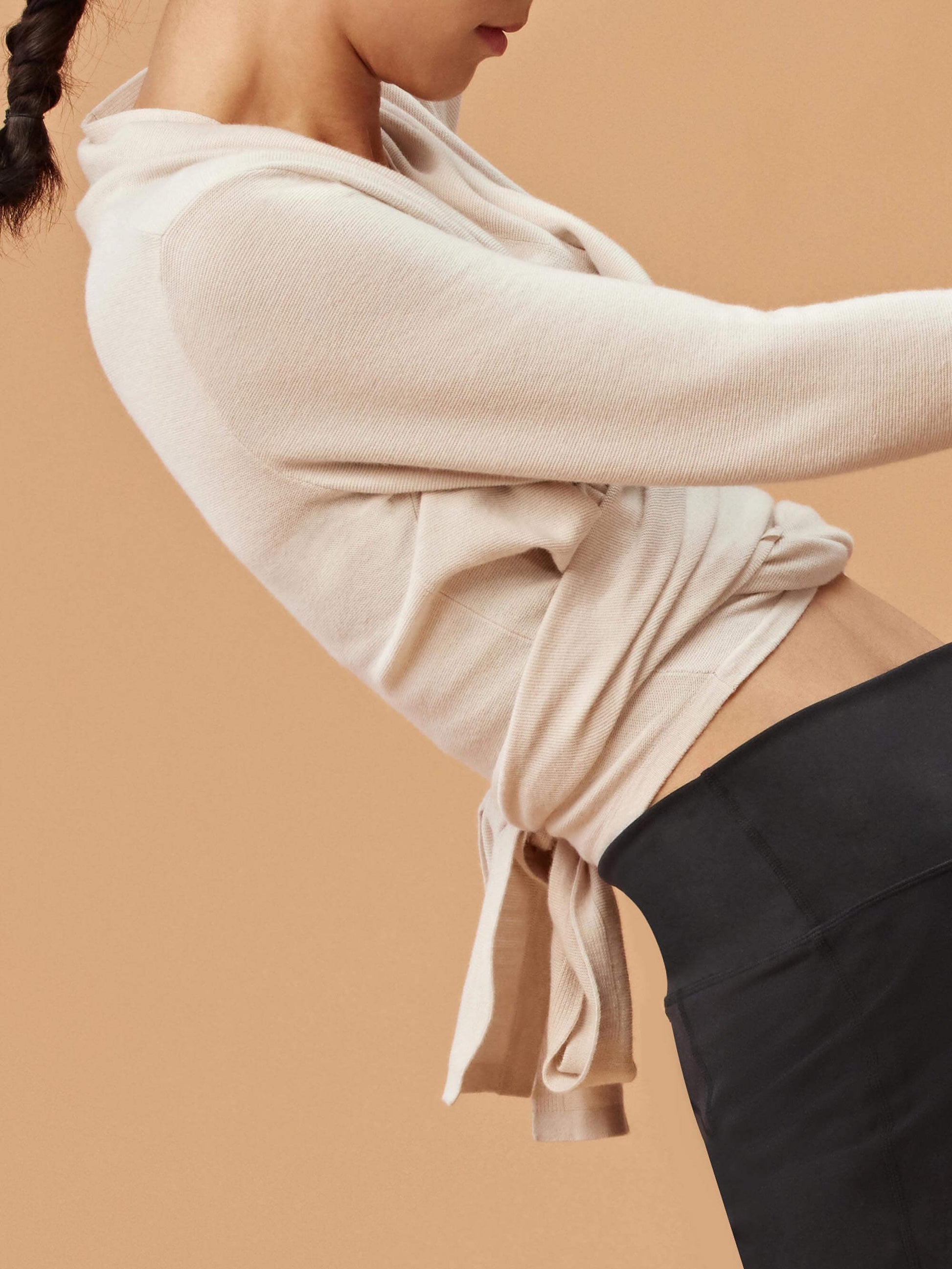 Close-up of a woman wearing a cream-colored wrap-style cardigan tied at the back, over a dark sports bra and high-waisted black pants. The cardigan has a soft, cozy texture, and the tied back detail adds a stylish touch.