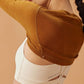 A close-up view of a woman wearing a white sports bra with the words “NEIWAI ACTIVE” printed on it and a brown ballet wool ribbed shrug. The woman has her arms raised, showing the detail of the shrug and sports bra, against a light brown background.