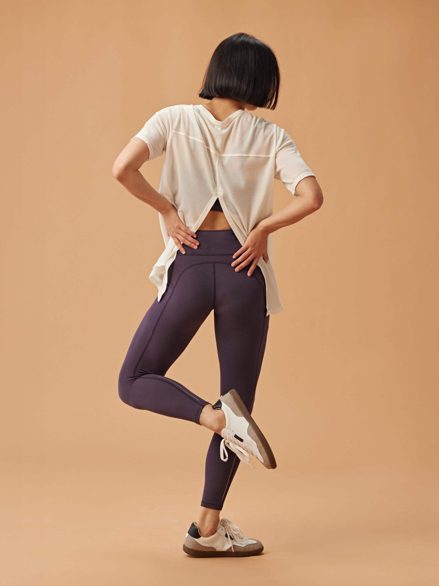 A woman wearing a light blue waffle short sleeve top with an open-back design, paired with dark purple leggings, posed with her back to the camera, one foot lifted, and hands resting on her hips.