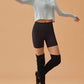 A woman in a light blue waffle long-sleeve top and black high-waisted shorts, striking a dance pose with one arm extended and one leg lifted, wearing black leg warmers and black ballet flats.