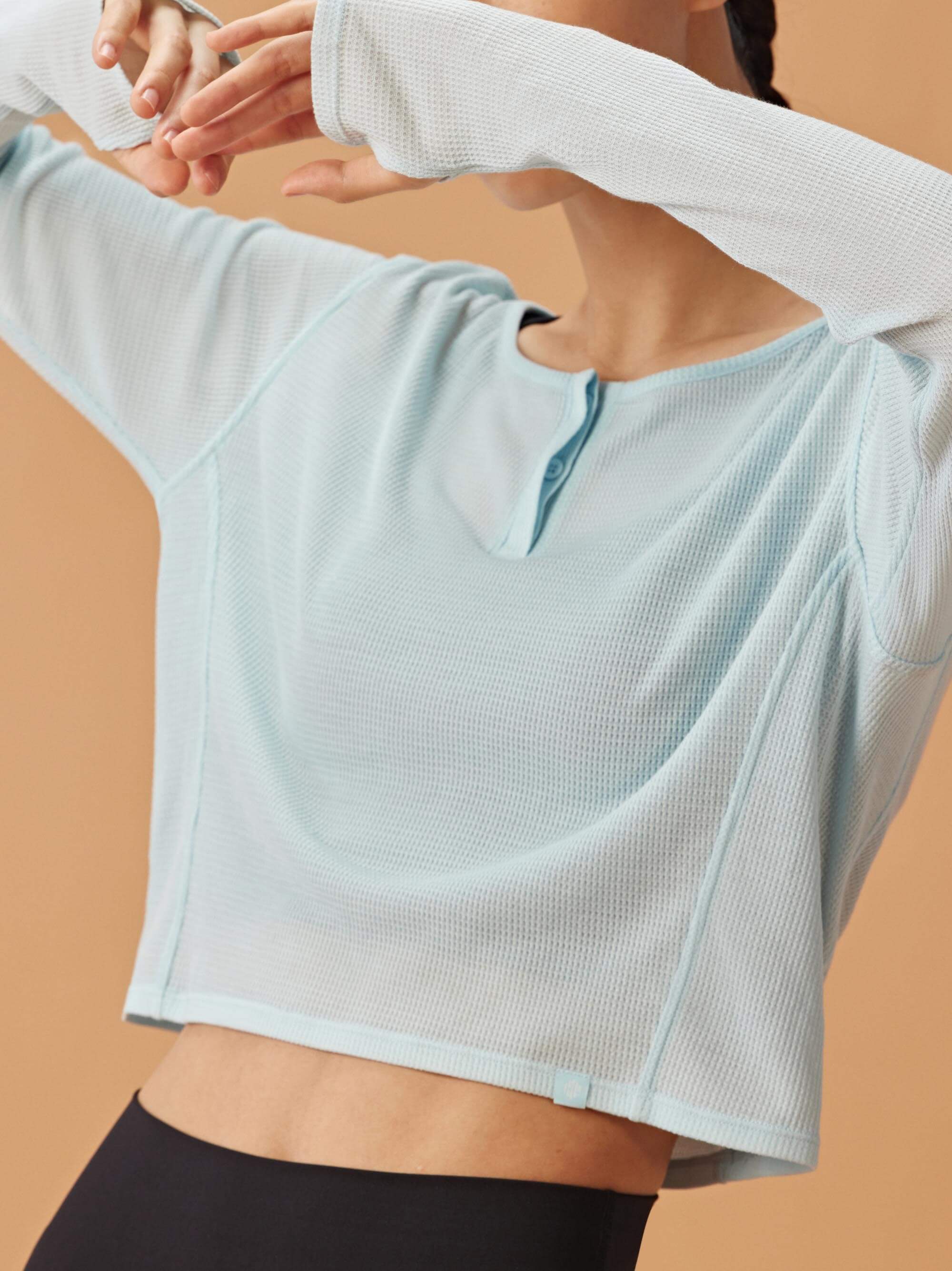A woman in a light blue, waffle long-sleeve top, covering her face with her arm, wearing black high-waisted shorts.