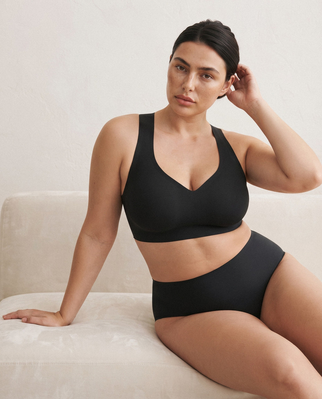 A woman wearing a black bralette and matching high-waisted briefs, seated on a light beige sofa against a neutral wall. black