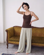 A woman stands barefoot in front of a vintage olive-green sofa, wearing a fitted brown camisole and loose light beige wide-leg pants. Seal Brown