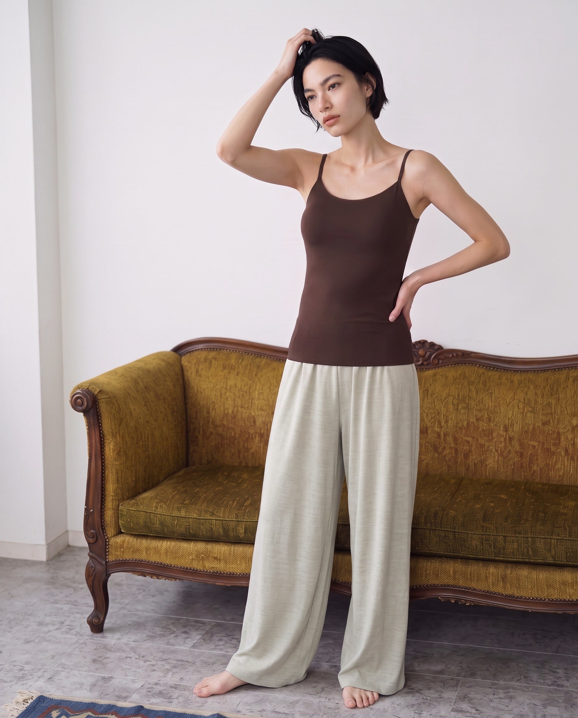 A woman stands barefoot in front of a vintage olive-green sofa, wearing a fitted brown camisole and loose light beige wide-leg pants. Seal Brown