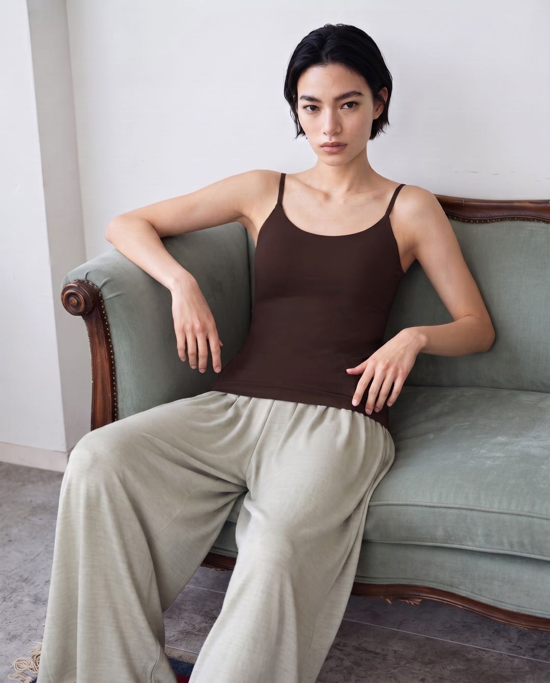 A woman sits on a muted green sofa wearing a dark brown spaghetti-strap camisole tank top and light beige wide-leg trousers in a minimalist indoor setting. Seal Brown