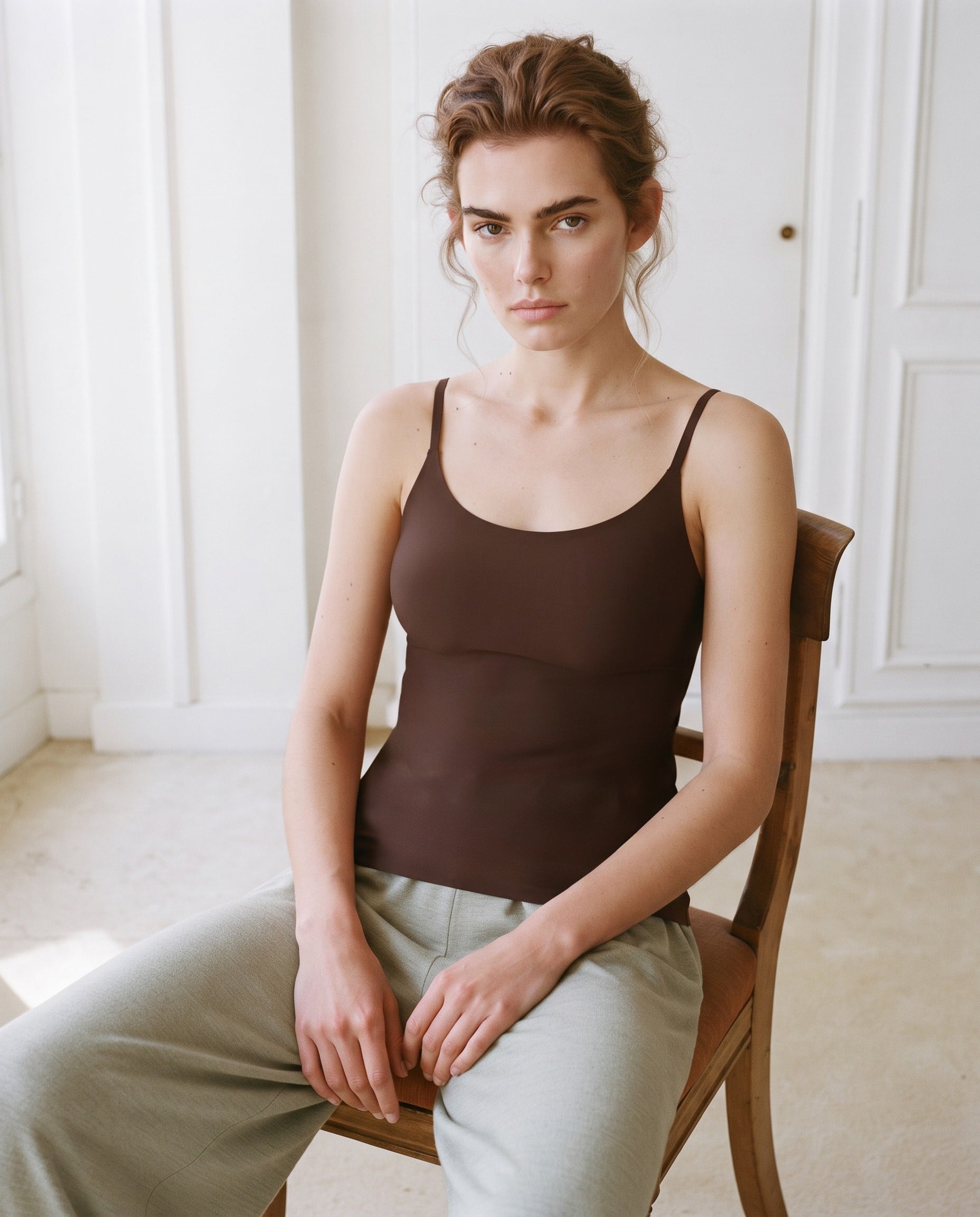 A woman sits on a wooden chair in a bright, white-paneled room, wearing a dark brown camisole tank top and light olive green trousers. Seal Brown