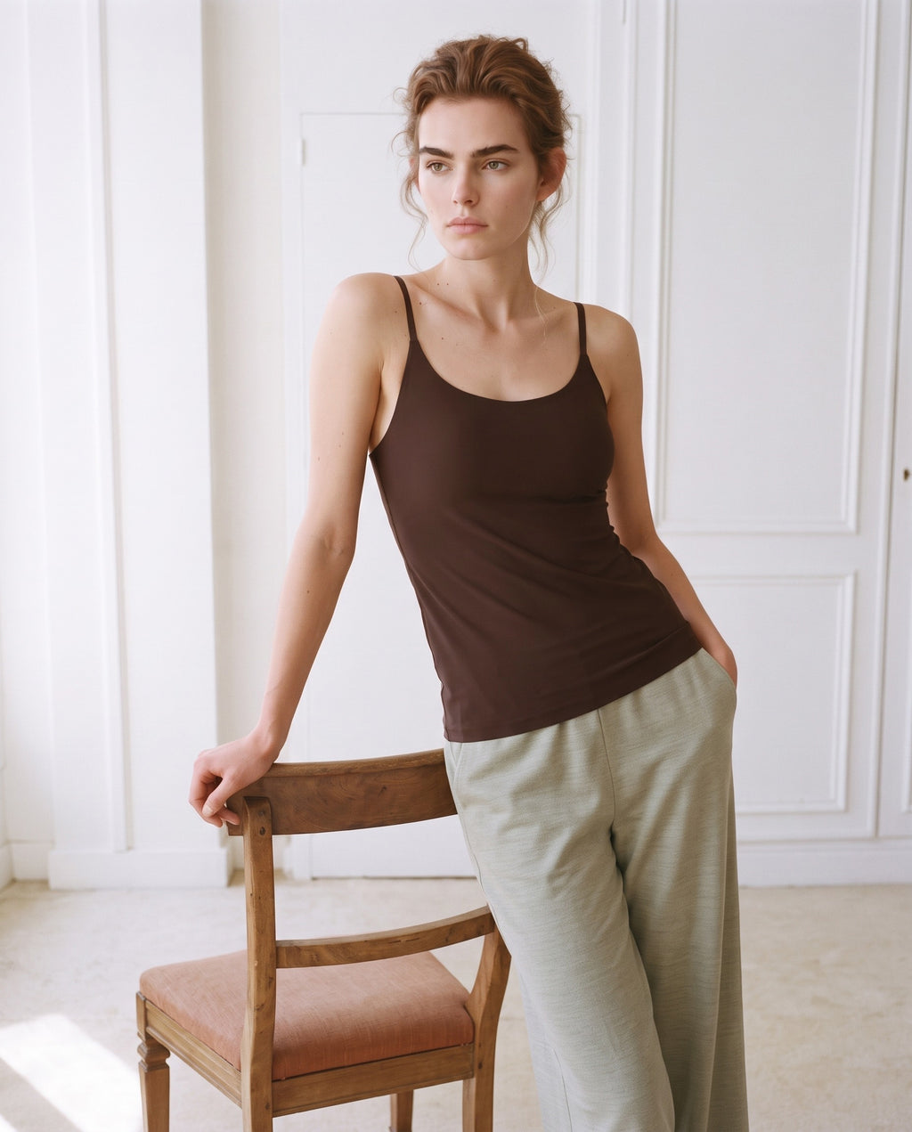 A woman wearing a dark brown ribbed tank top and light olive green wide-leg linen trousers, leaning against a wooden chair in a bright, white-paneled room. Seal Brown