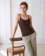 A woman wearing a dark brown ribbed tank top and light olive green wide-leg linen trousers, leaning against a wooden chair in a bright, white-paneled room. Seal Brown