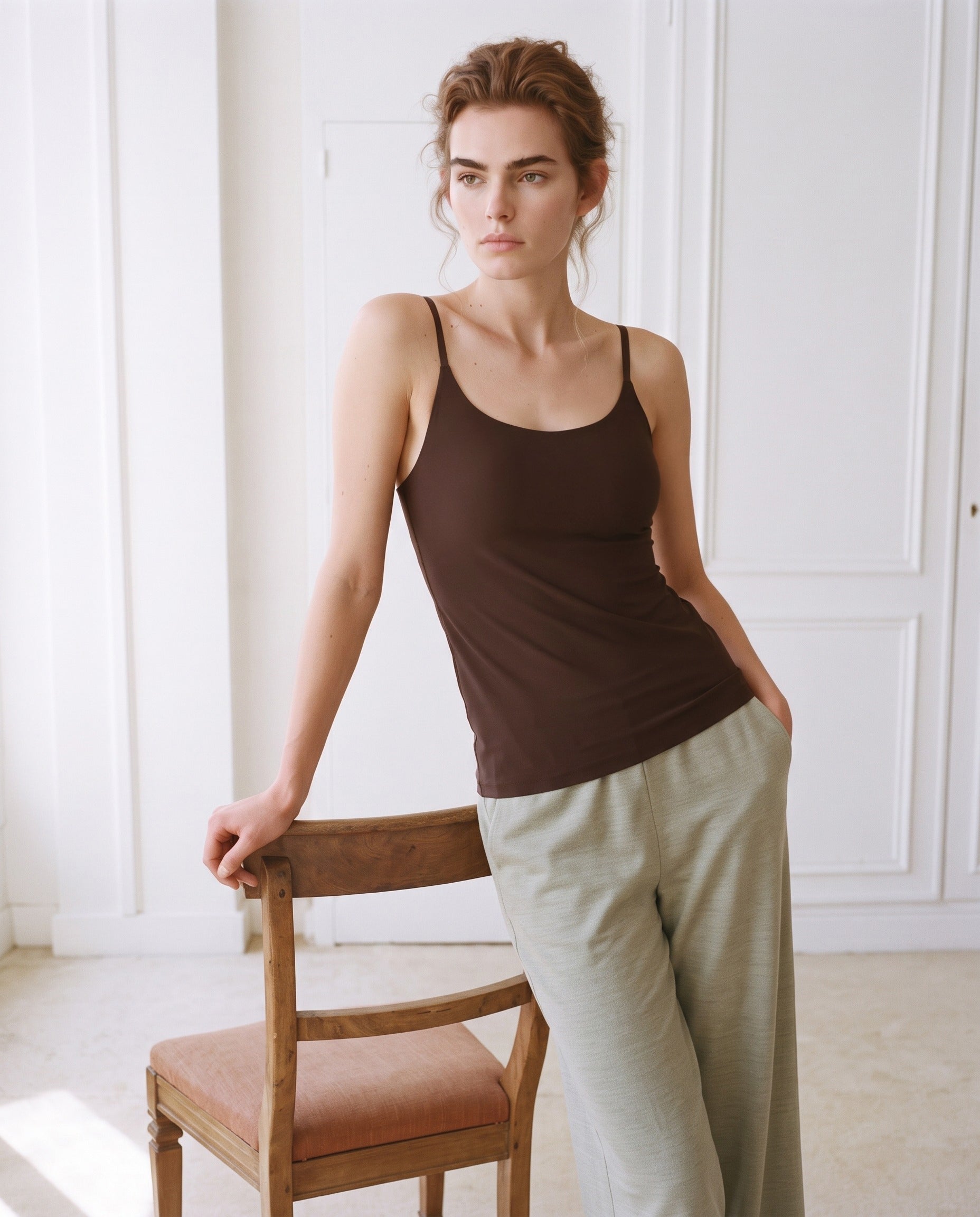 A woman wearing a dark brown ribbed tank top and light olive green wide-leg linen trousers, leaning against a wooden chair in a bright, white-paneled room. Seal Brown