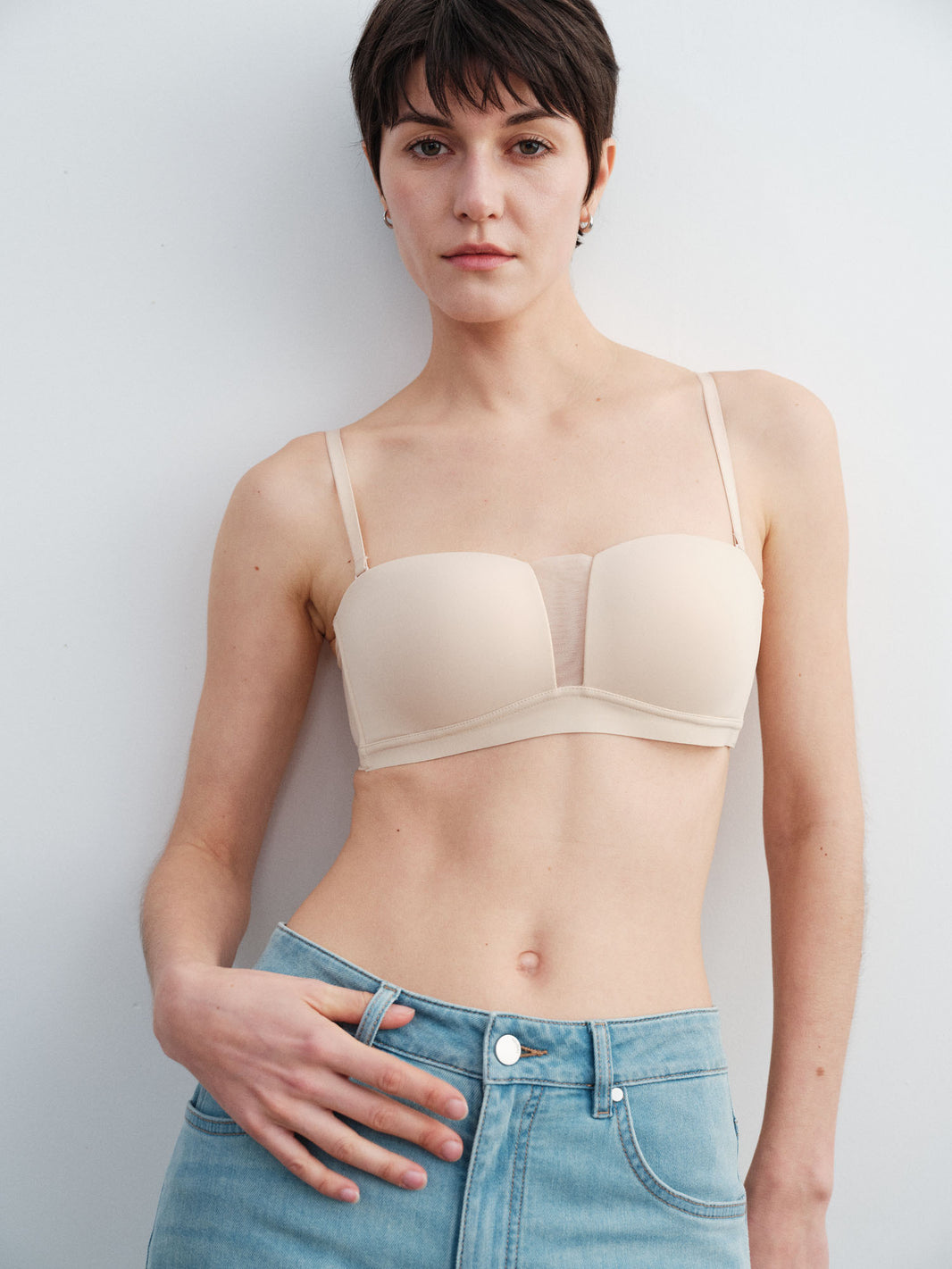 Model wearing a beige bra and blue jeans against a plain background.