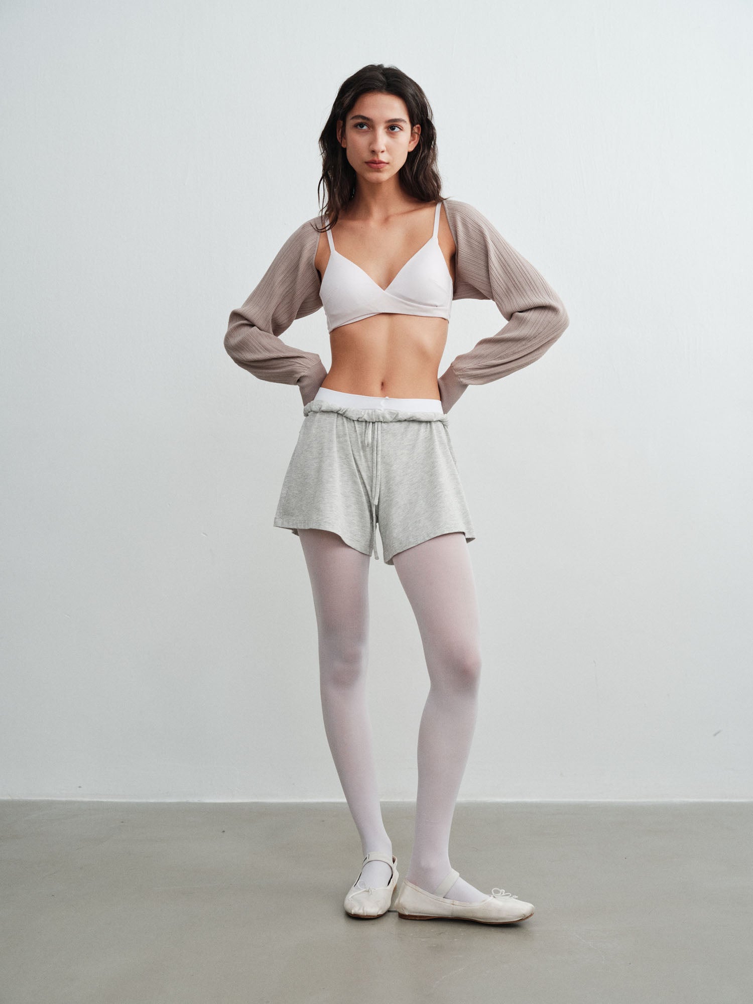 Model wearing a white sports bra and shorts with a gray cardigan on a plain background.