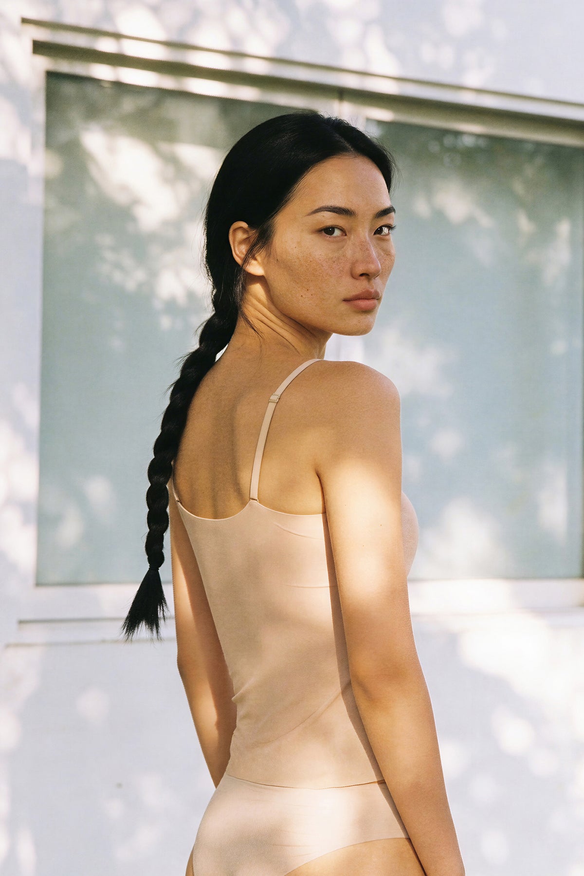 An Asian woman with freckles and a long dark braid looks back over her shoulder, wearing a nude beige camisole bralette and matching bottoms, dappled sunlight falling across her back against a pale wall.