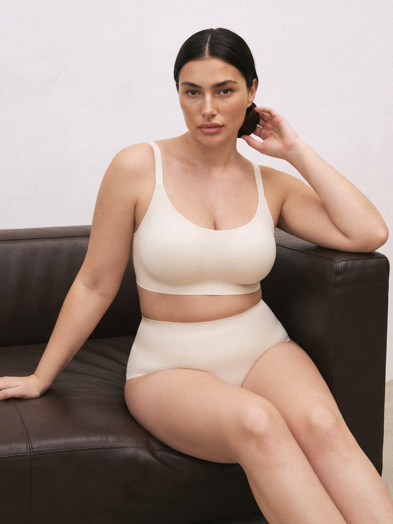 A woman sits on a dark brown leather couch wearing a matching beige two-piece bra and panty set against a plain off-white wall. Vanilla Bean