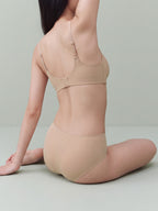 A woman wearing a matching beige bra and high-waisted brief set, shown from the back while kneeling on a light gray studio floor. Treacly Almond