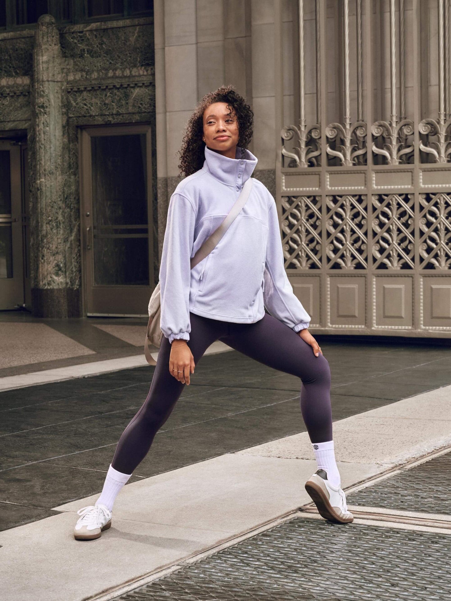 Woman wears light purple sweatshirt, dark purple leggings and gray crossbody bag, standing and pointing one foot.