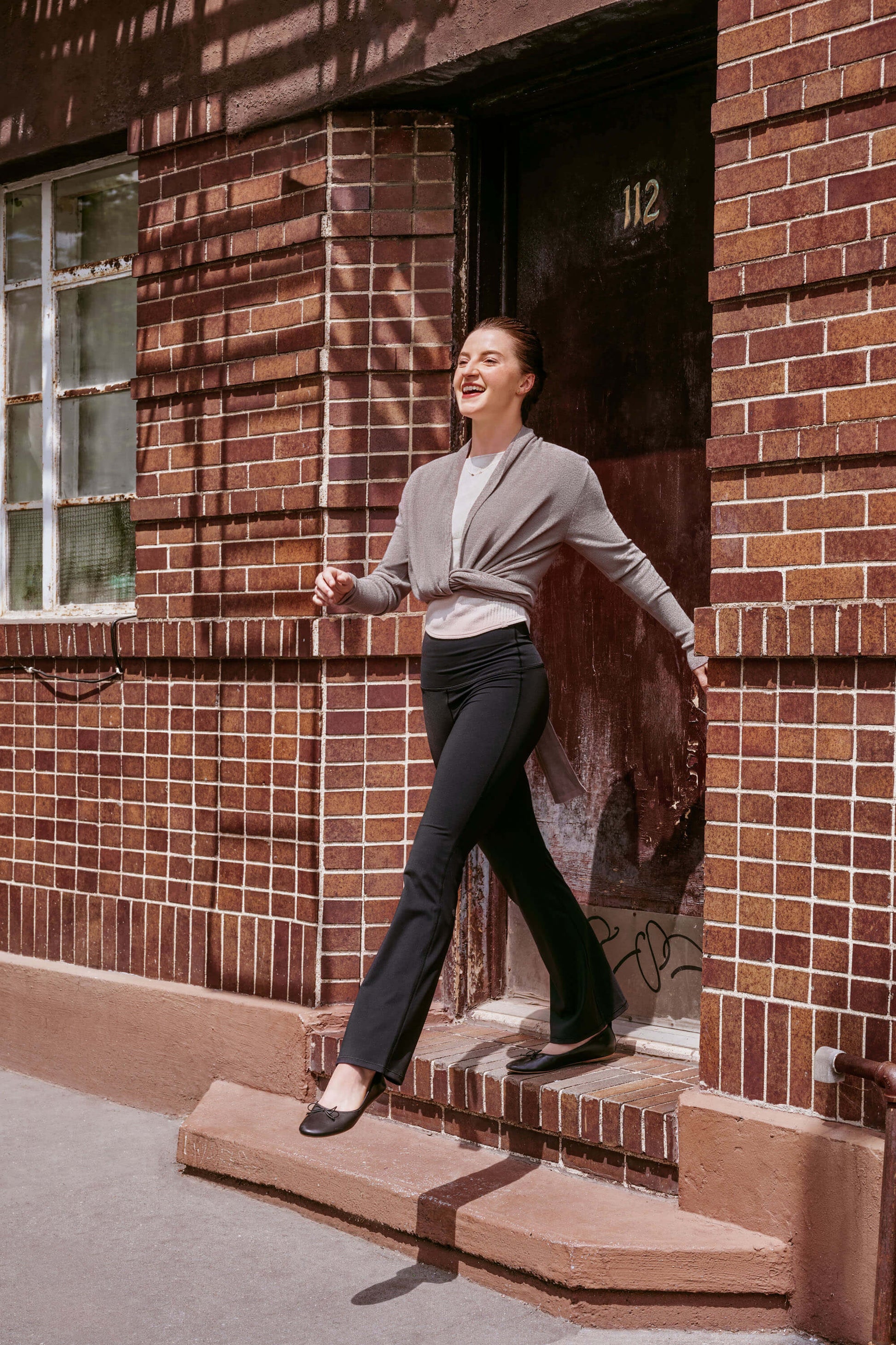 Dancer stepping from doorway wearing gray wrap sweater, white tank top, and black legging pants