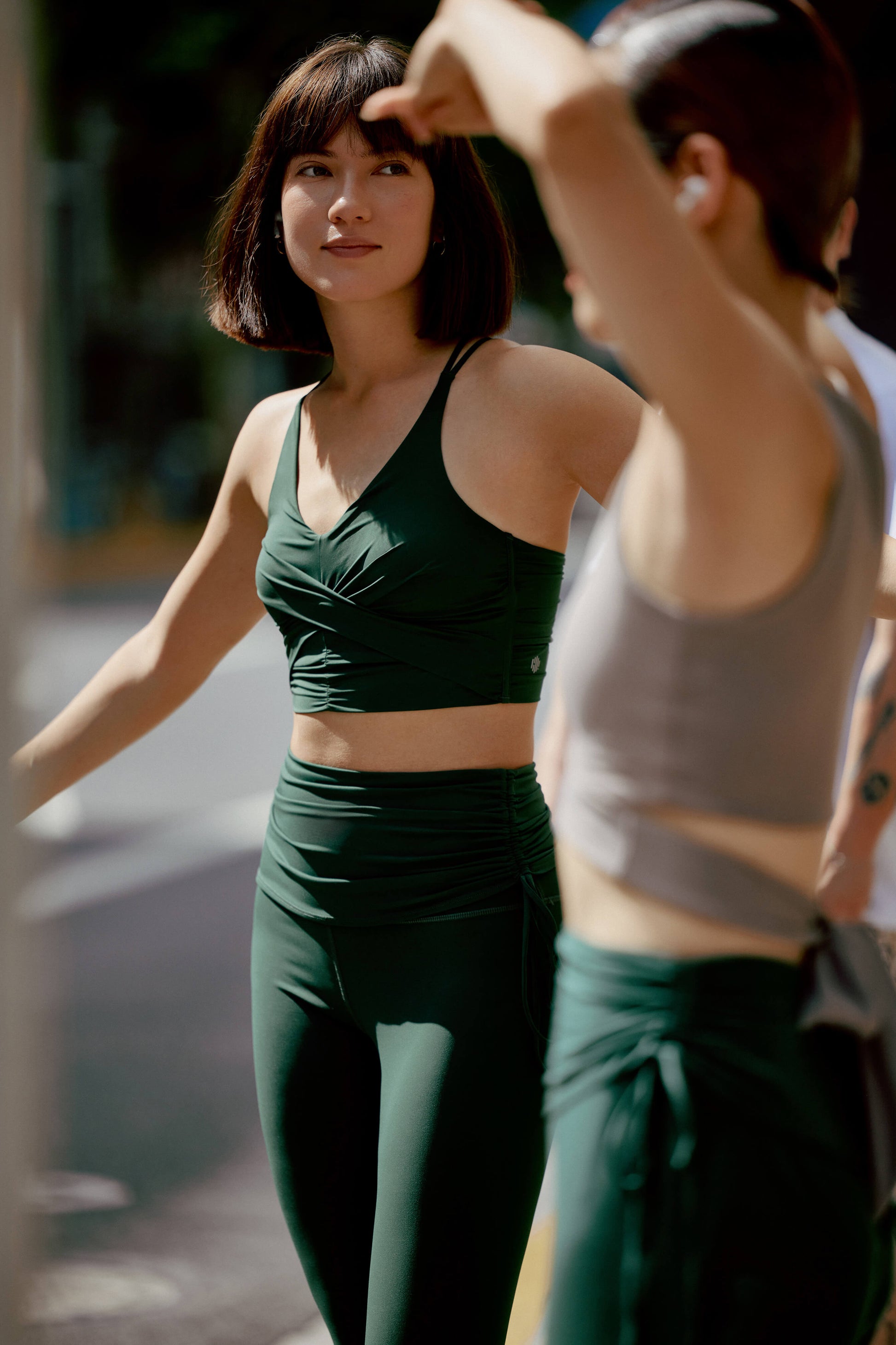 two women in workout outfit. One facing front is wearing a dark green v-neck sports bra with matching double layered waist leggings with side knots. One woman facing side is wearing a light grey top and same dark green leggings as the other woman.
