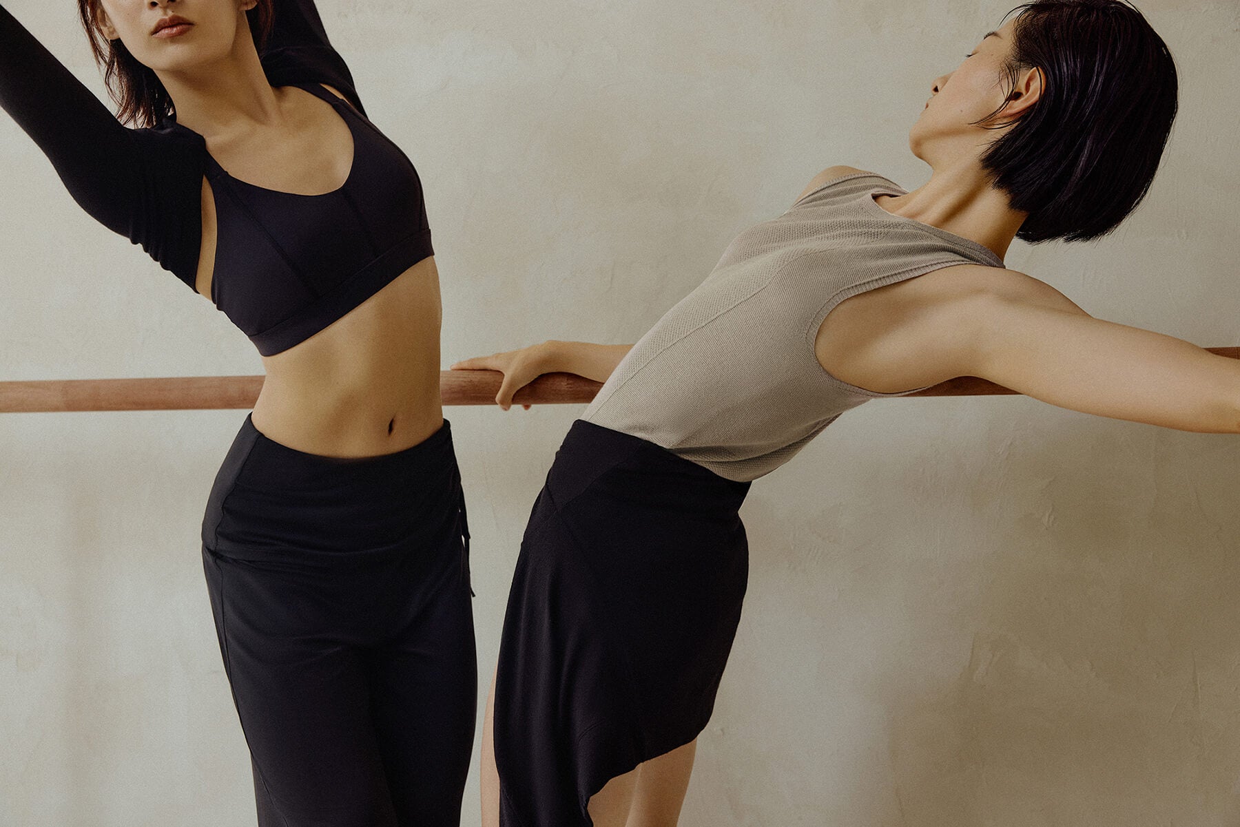 Two women at barre, one woman wearing black bra, skirt and shrug, the other woman wearing gray tank top and black skirt