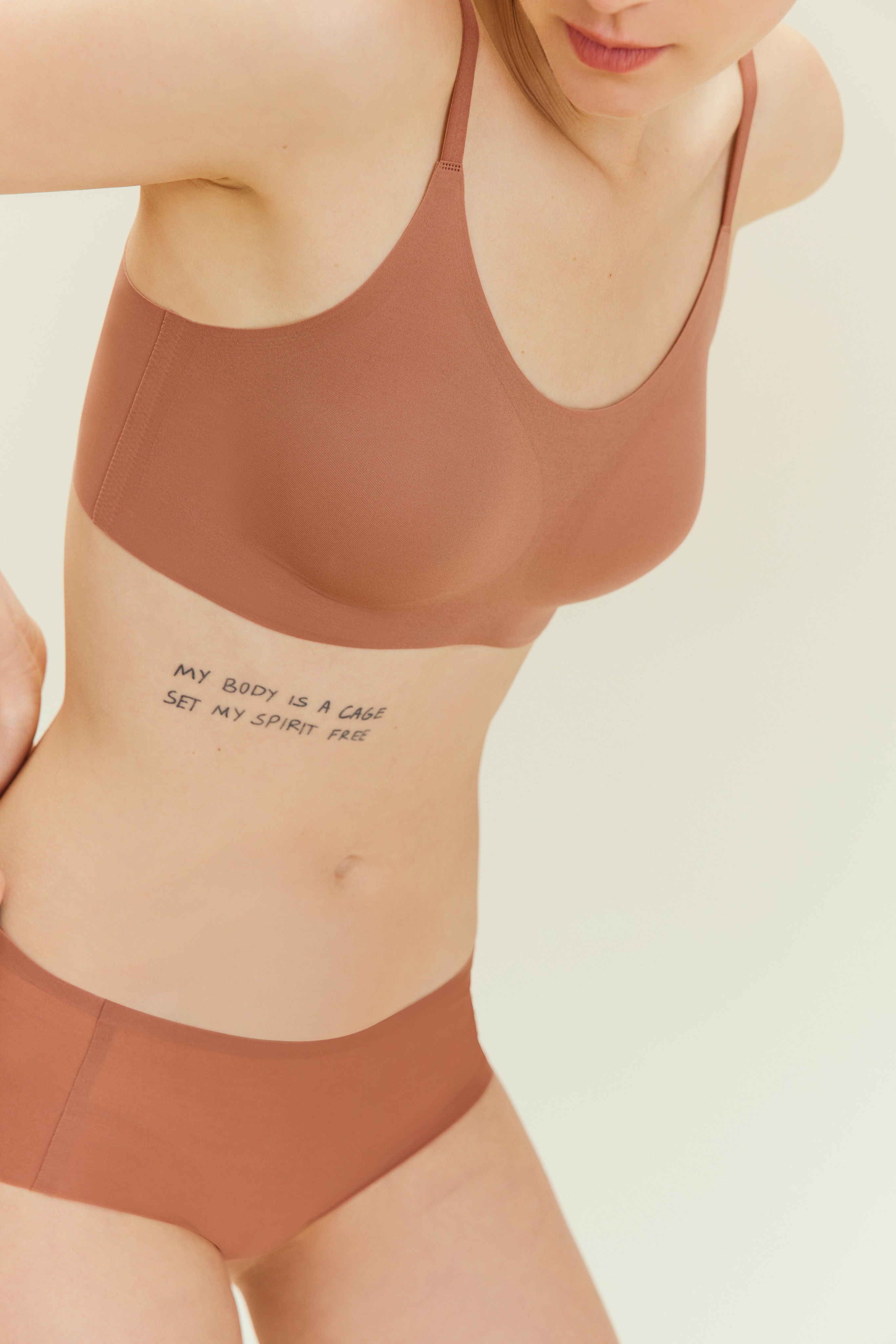 Person wearing a brown bra and underwear set with a visible tattoo on a plain background