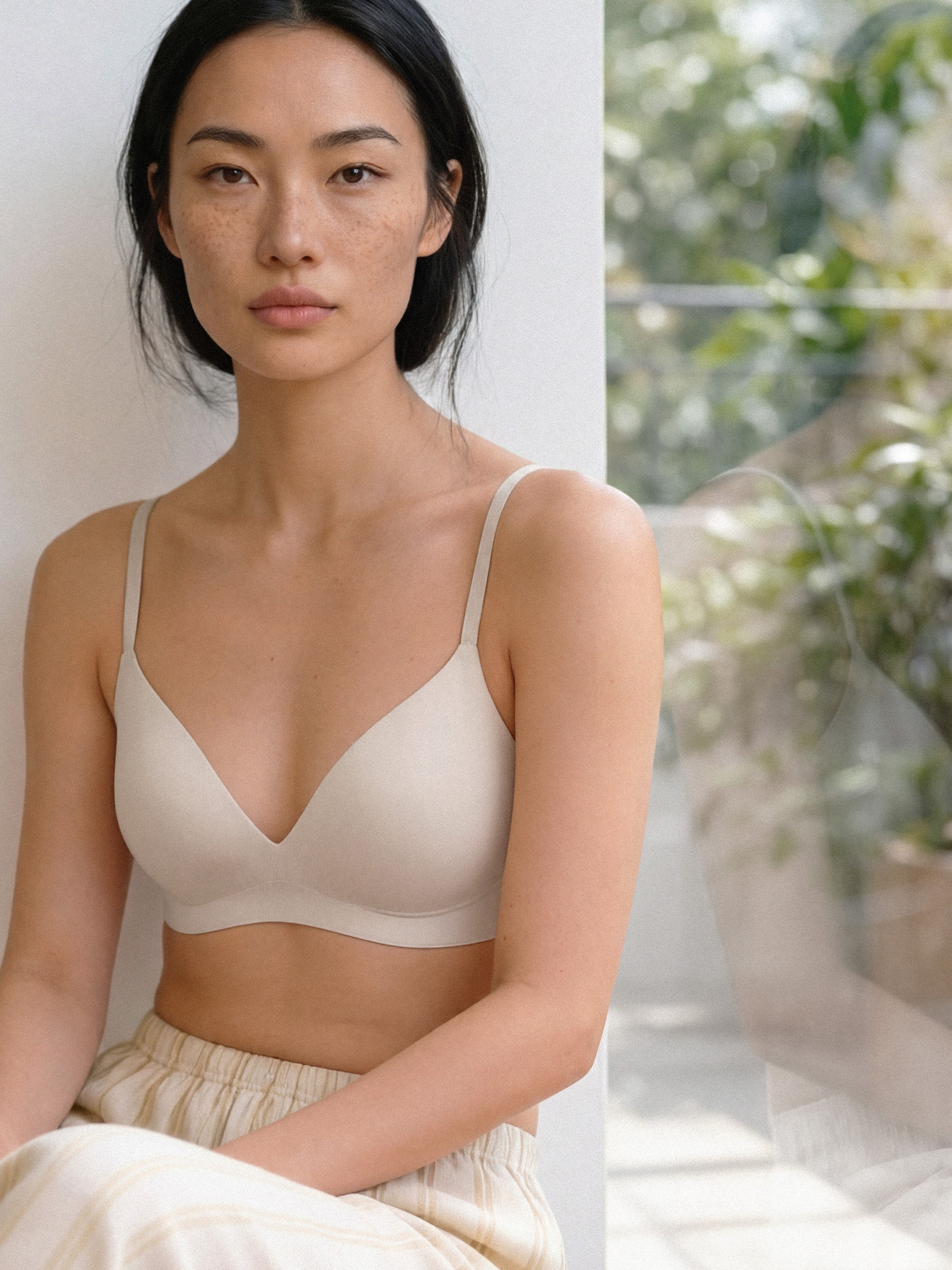 Woman wearing a beige bralette in a softly lit room with plants in the background