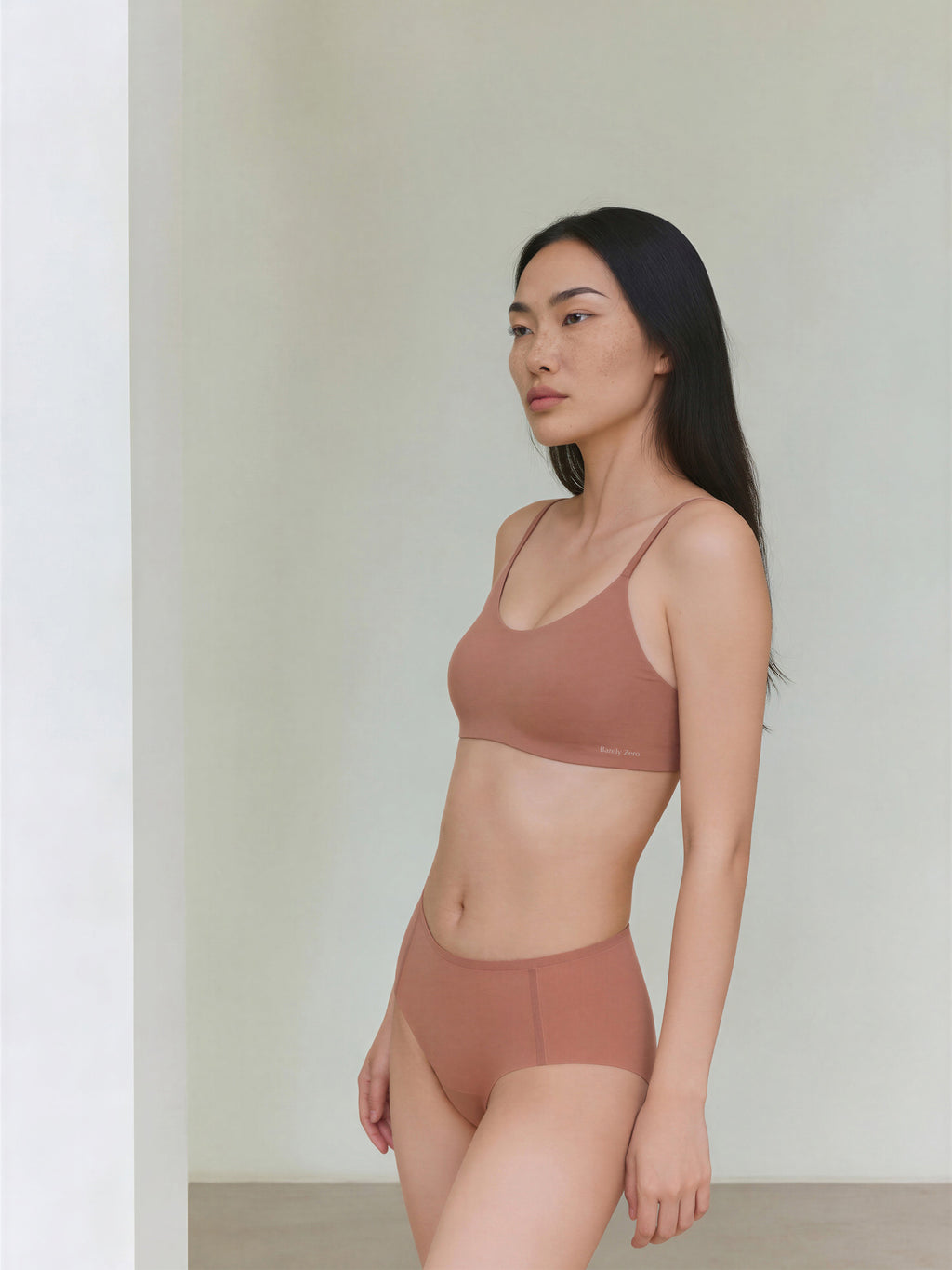 A woman with long dark hair stands indoors, wearing a Barely Zero Strap Bra and matching light brown underwear. She faces left with a neutral expression, showcasing invisible comfort against a minimal, light background. Cinnamon Taste color.