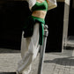 A woman wearing a white sports cardigan, green sports bra, and white sports pants leans on the railing. There is a building in the background.