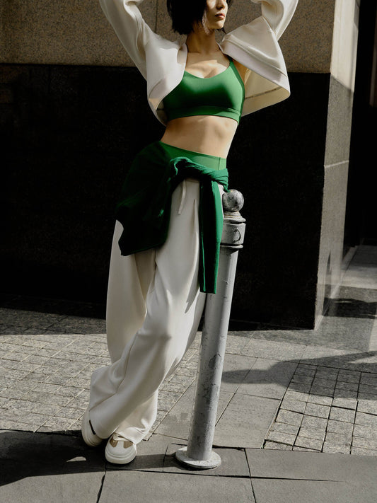 A woman wearing a white sports cardigan, green sports bra, and white sports pants leans on the railing. There is a building in the background.