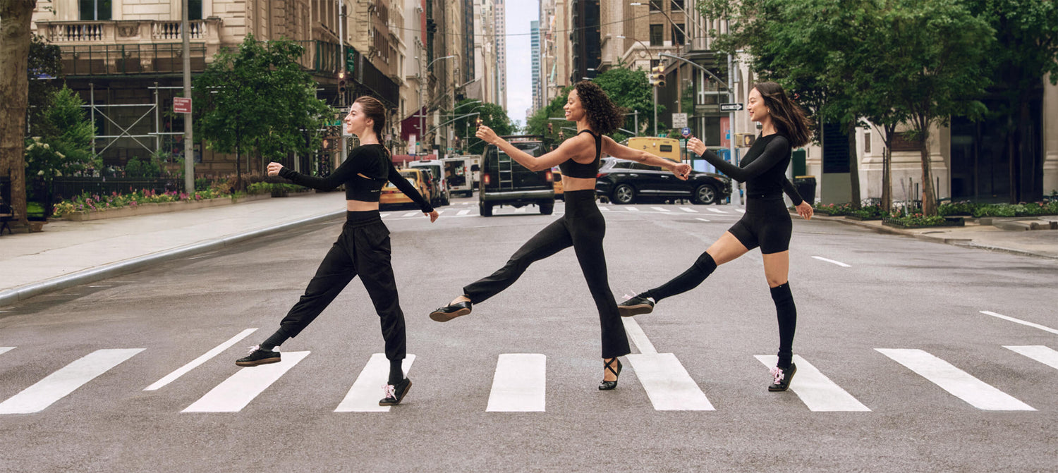 Three dancers wearing black walking on crosswalk with one leg kicked out in front