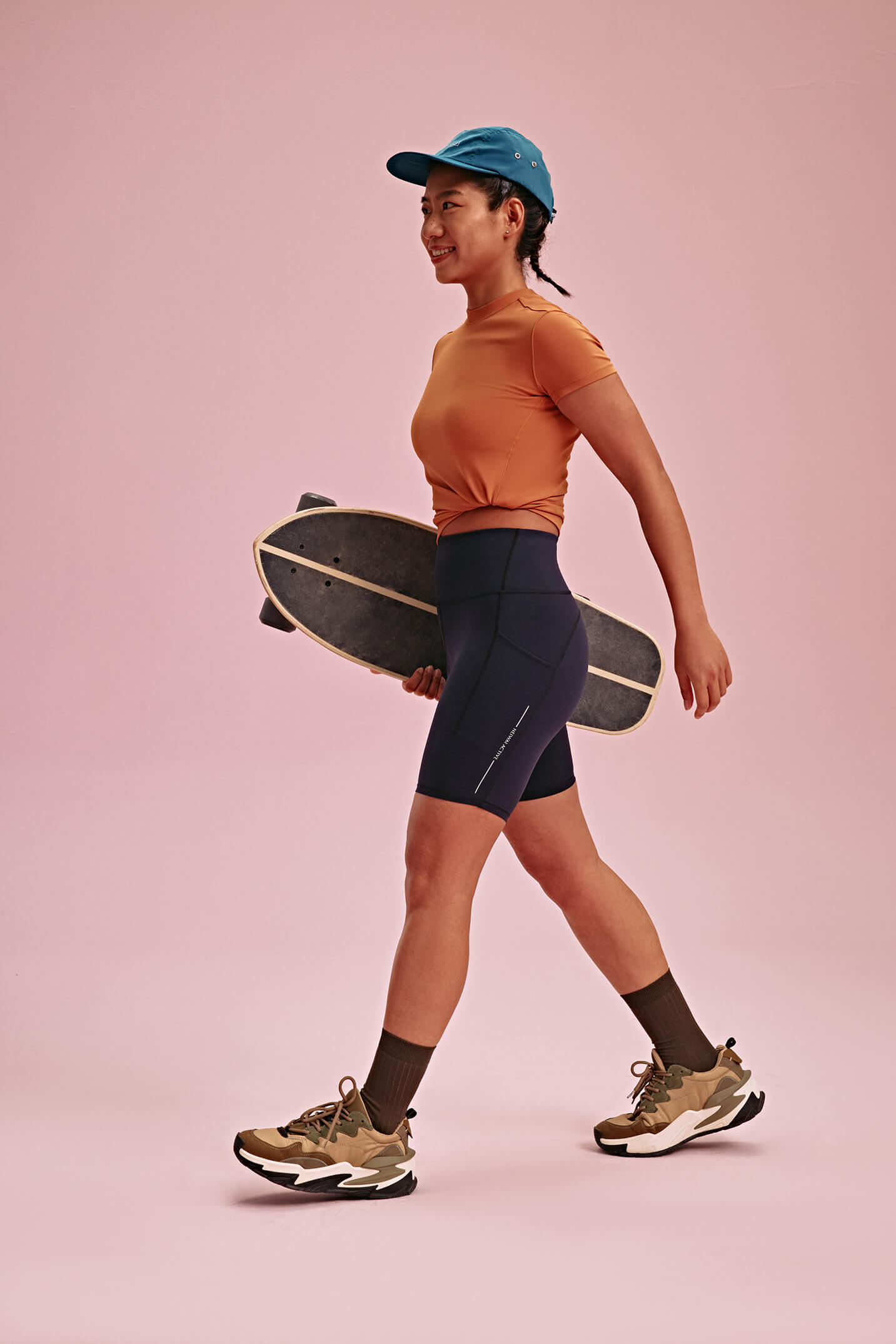 Woman wearing orange T-shirt and black biker shorts carrying a longboard